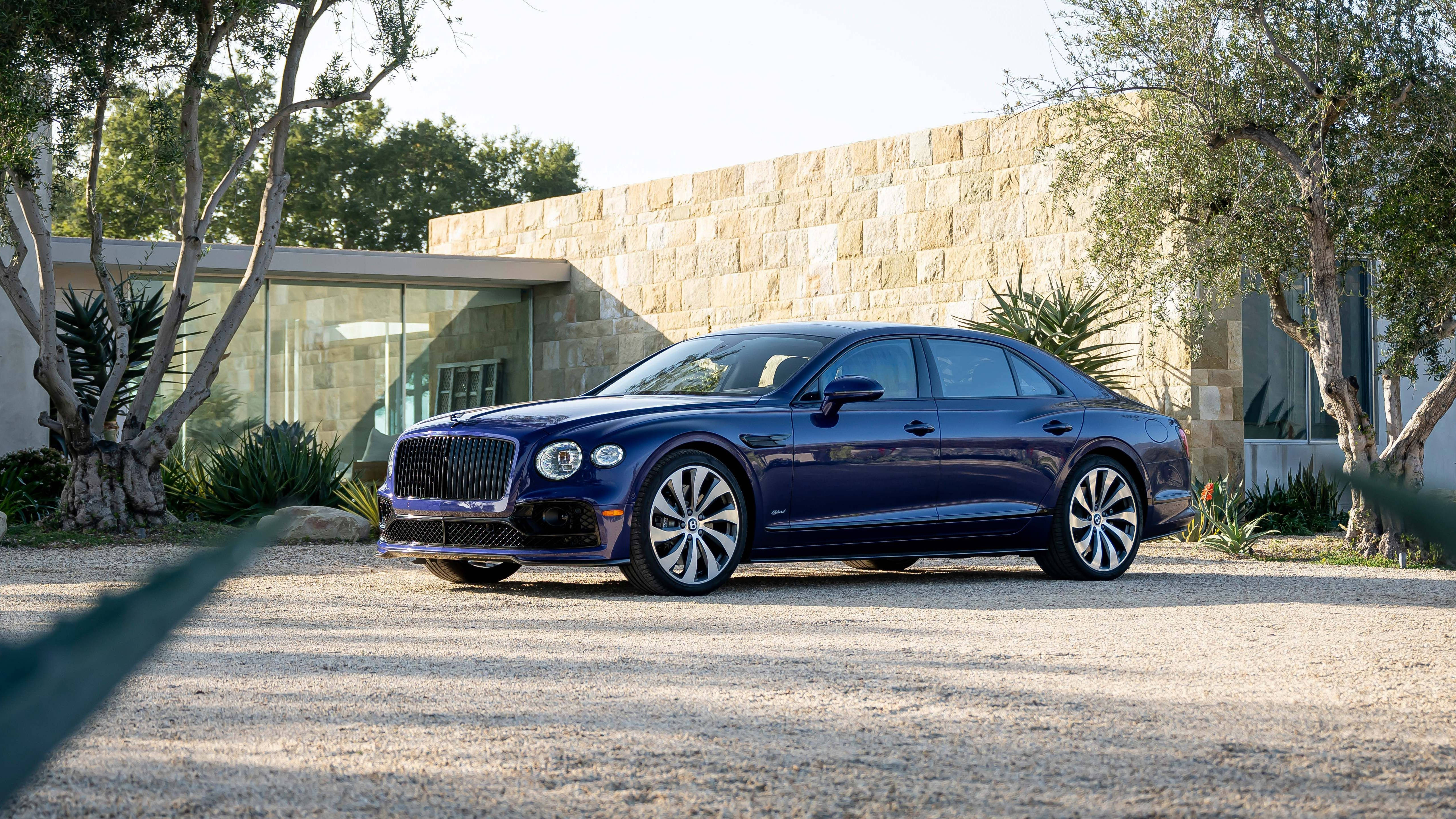 4K 桌布 壁紙 — 一輛時尚的深藍色 Bentley Flying Spur 停泊在碎石車道上，前面是一座被樹木環繞的現代石頭和玻璃房屋。