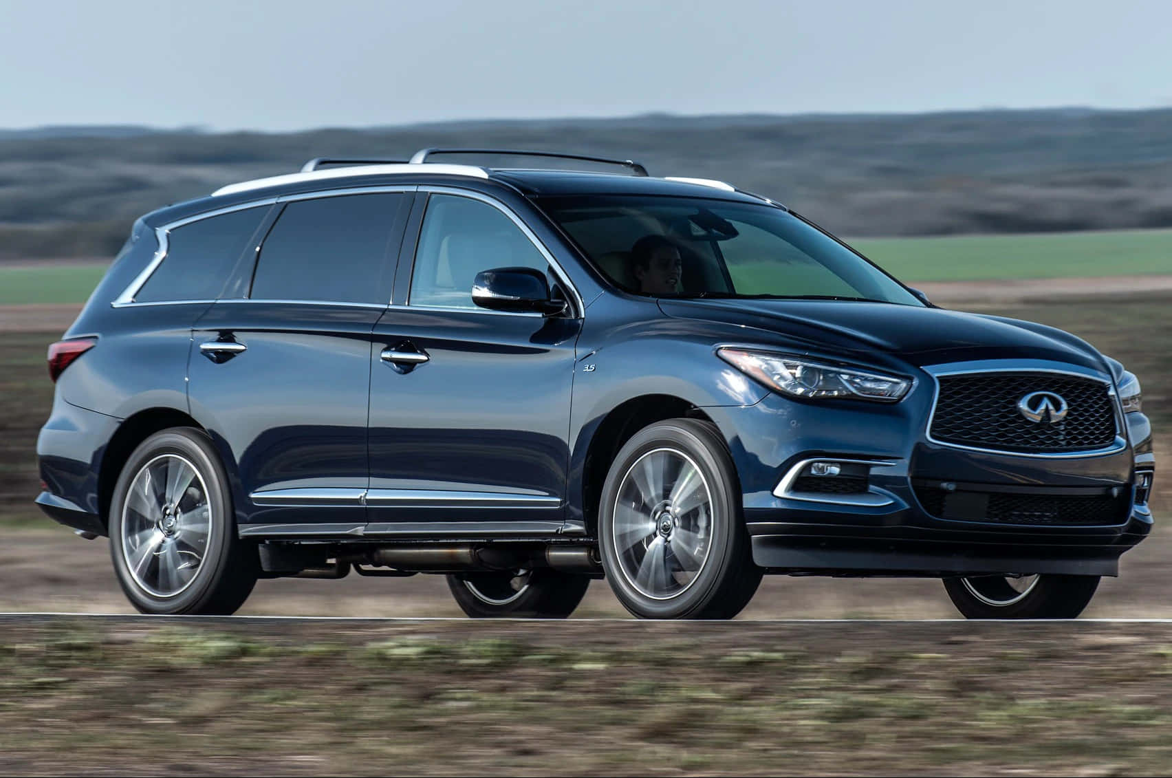 Infiniti Qx60 桌布 壁紙 — 一輛時尚的深藍色 Infiniti QX60 SUV 在鄉間小路上行駛，背景是模糊的草地和遠處的山丘，天空晴朗。