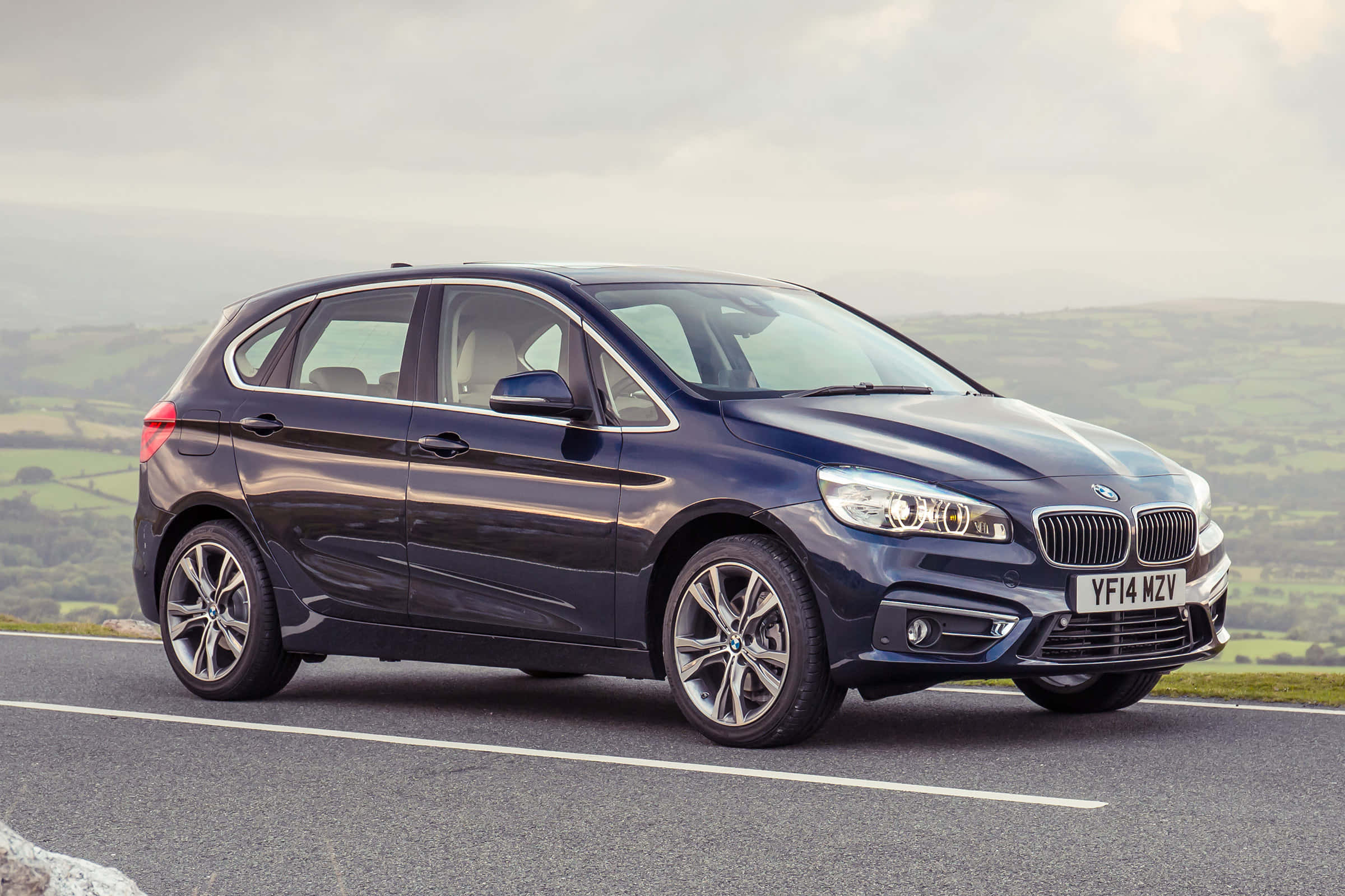 Bmw 2 Series 桌布 壁紙 — 一輛時尚的深藍色BMW 2系列Active Tourer在陰天下行駛在蜿蜒的鄉村道路上，兩旁是起伏的綠色山丘。