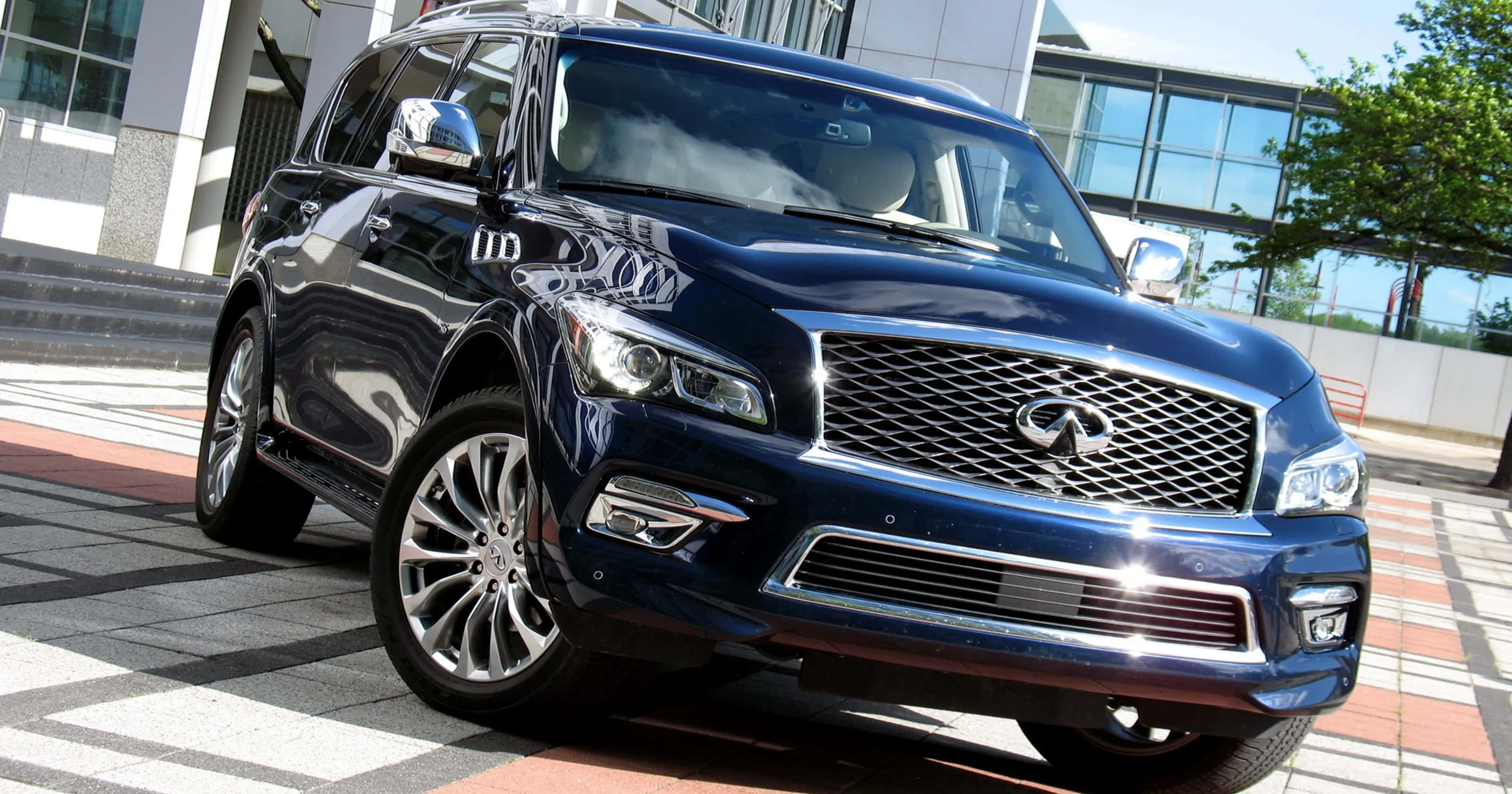 Infiniti Qx80 桌布 壁紙 — 一輛時尚的深藍色Infiniti QX80 SUV在明亮的陽光下閃閃發光，停放在現代瓷磚廣場上，背景是玻璃建築。