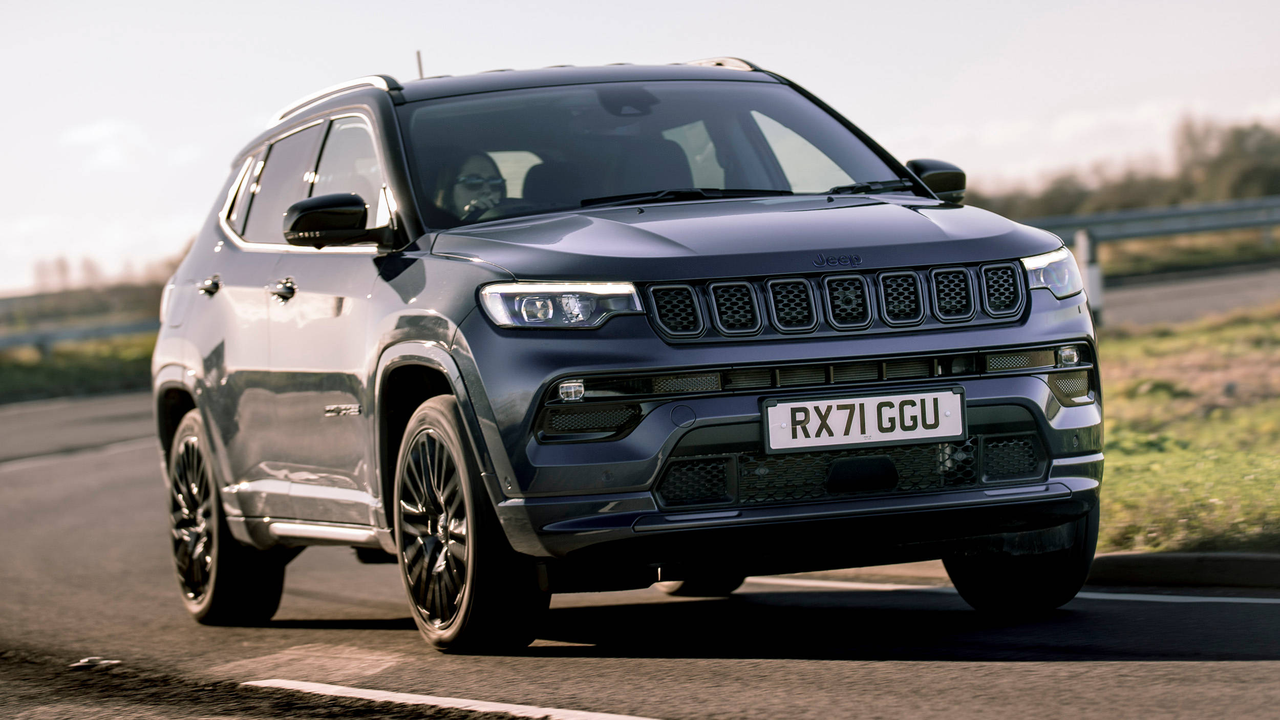 Road 桌布 壁紙 — 一輛時尚的深藍色Jeep Compass SUV在柔和日光下自信地行駛在蜿蜒的道路上，兩旁有草地和護欄。