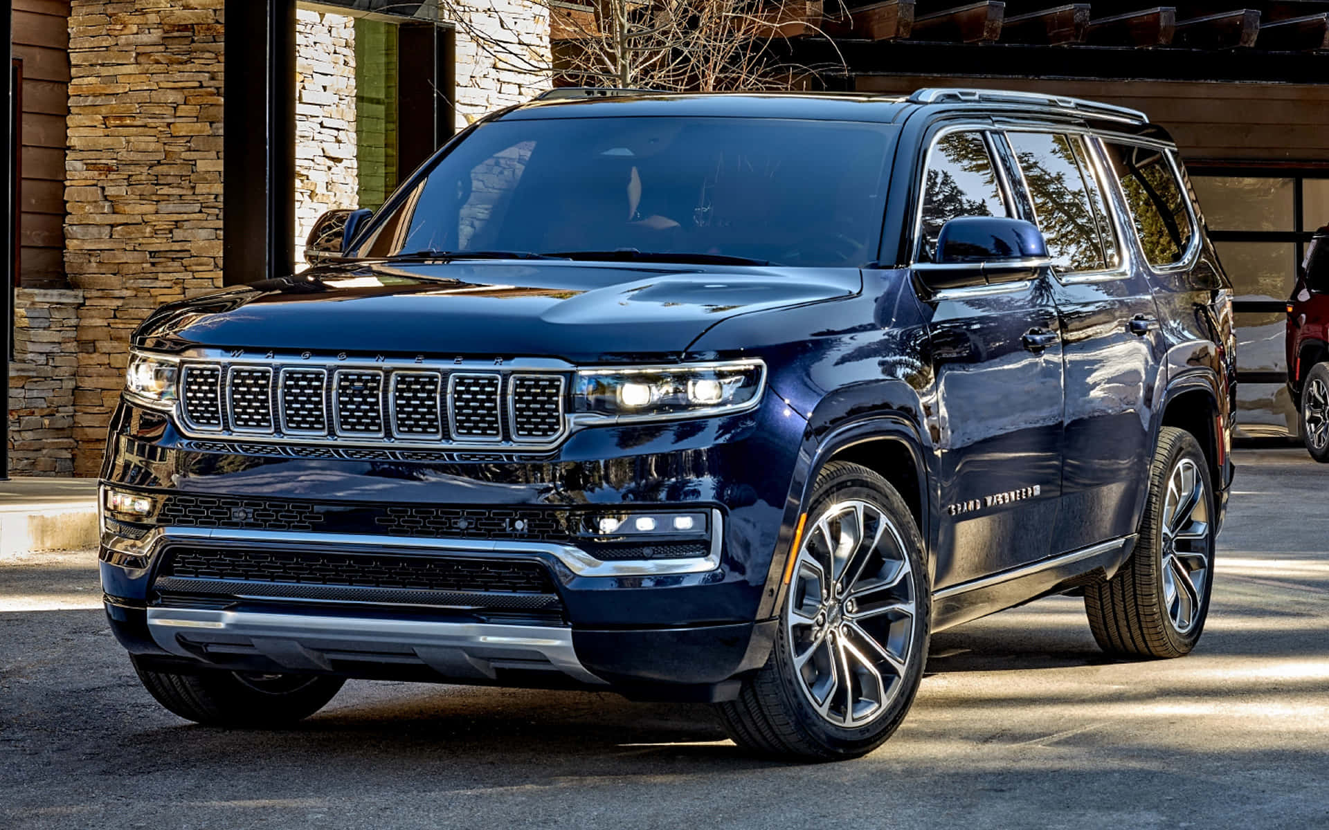 Jeep Wagoneer 桌布 壁紙 — 一輛時尚的深藍色Jeep Grand Wagoneer SUV在明亮的陽光下閃閃發光，停在一座石頭建築旁邊。