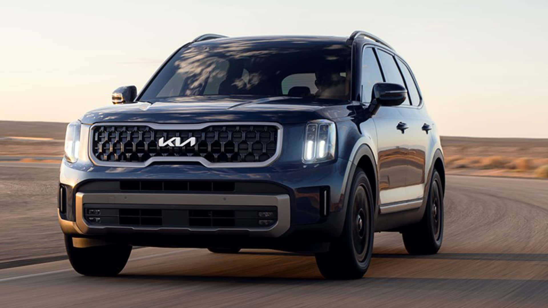 Kia Telluride 桌布 壁紙 — 一輛時尚的深藍色Kia SUV在柔和的金色日落天空下自信地行駛在沙漠公路上。