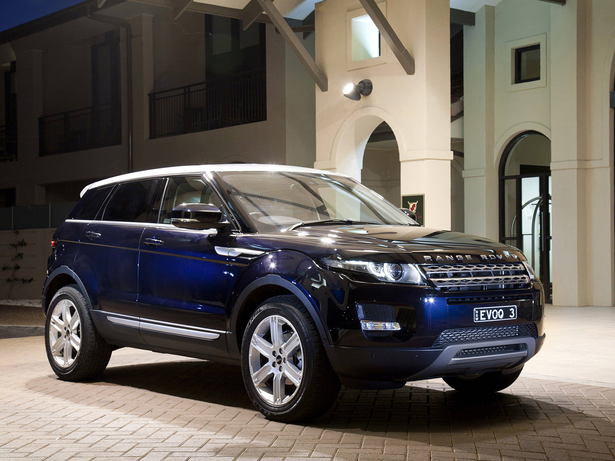 land rover iPhone 桌布 壁紙 — 一輛時尚的深藍色Range Rover Evoque在夜間停在一棟帶有拱形門廊的現代建築前。