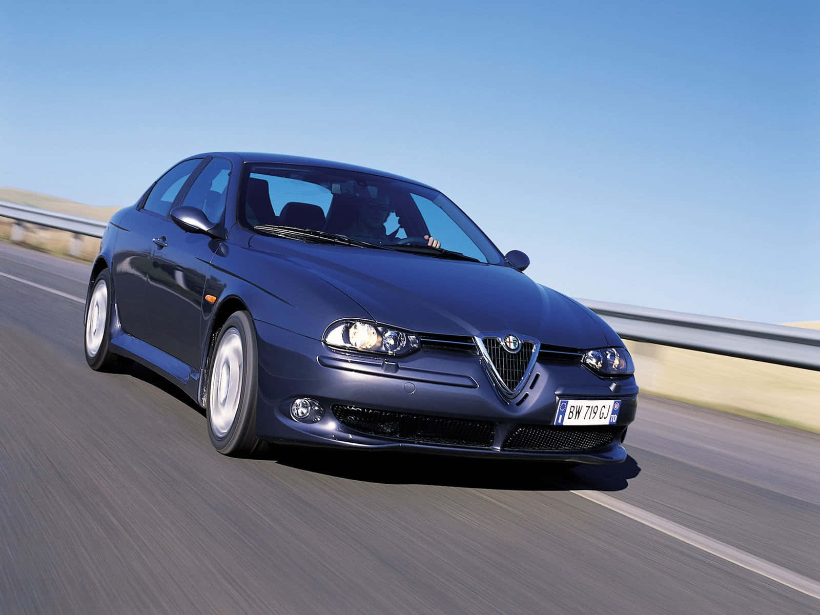 Alfa Romeo 156 桌布 壁紙 — 一輛時尚的深藍色愛快羅密歐跑車在晴朗的藍天下，沿著陽光普照的高速公路飛馳。