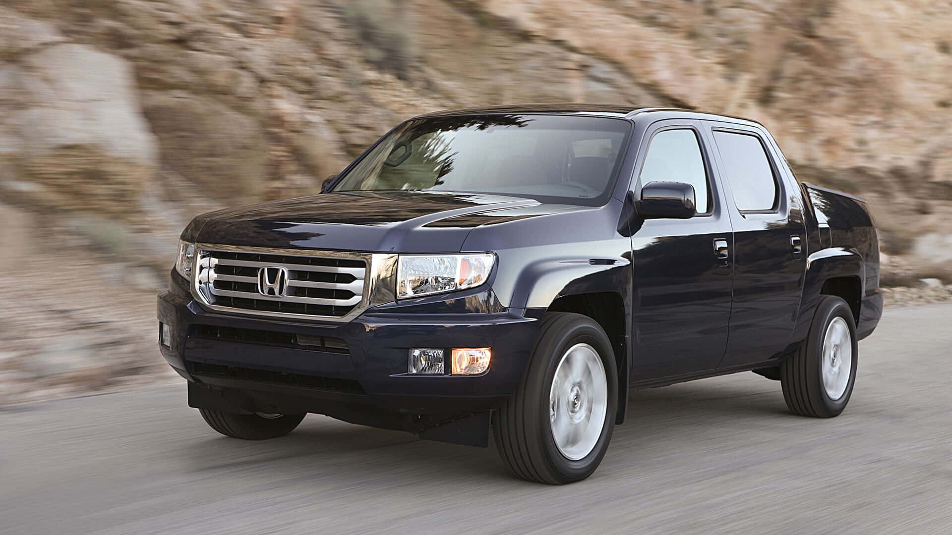 Honda Ridgeline 桌布 壁紙 — 一輛時尚的深藍色本田Ridgeline皮卡車沿著蜿蜒的山路疾馳，背景是模糊的岩石地形。