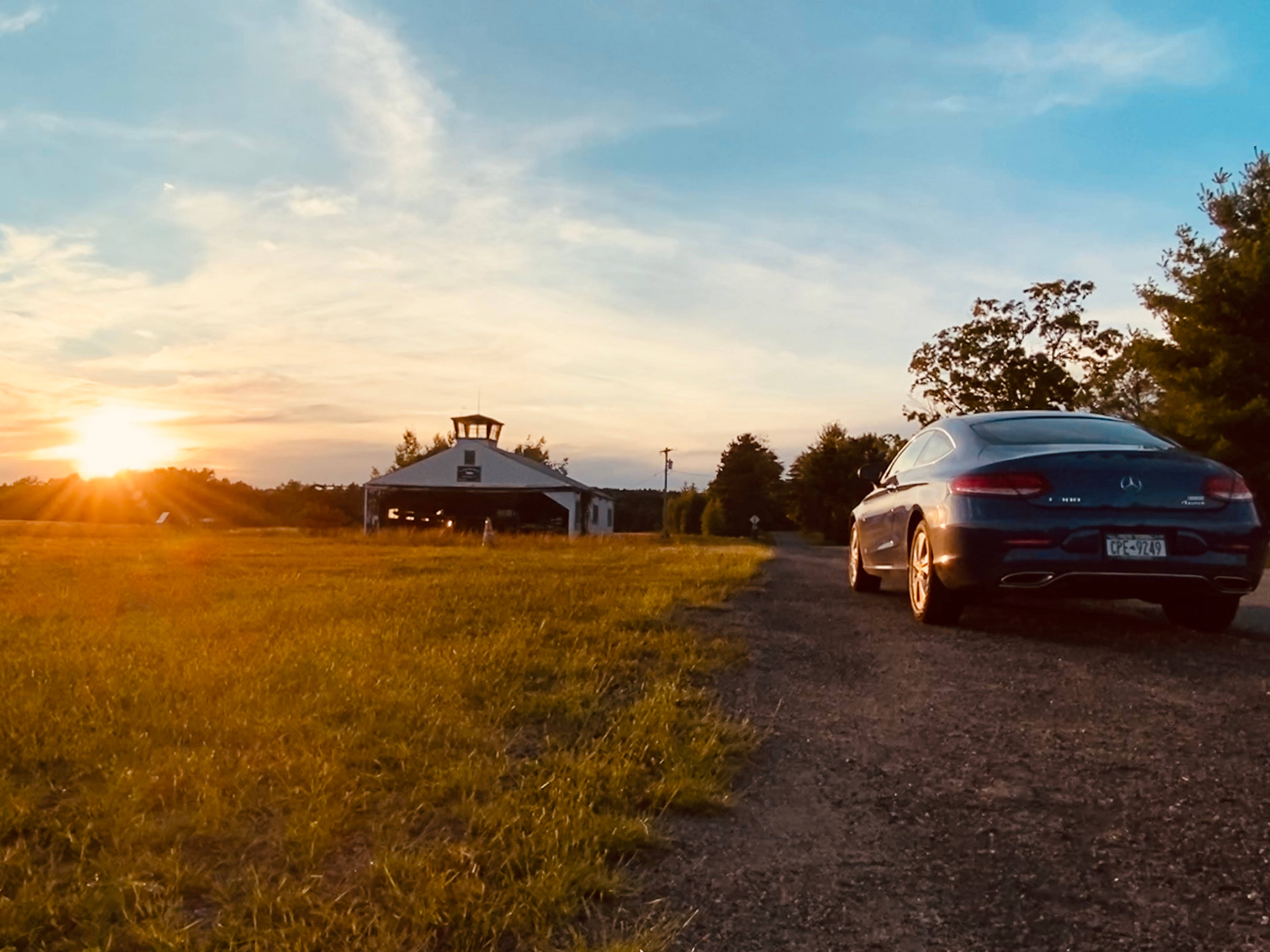 Mercedes Amg 桌布 壁紙 — 一輛時尚的深藍色汽車停在日落時分的碎石路上，旁邊是草地，遠處有一座白色穀倉。