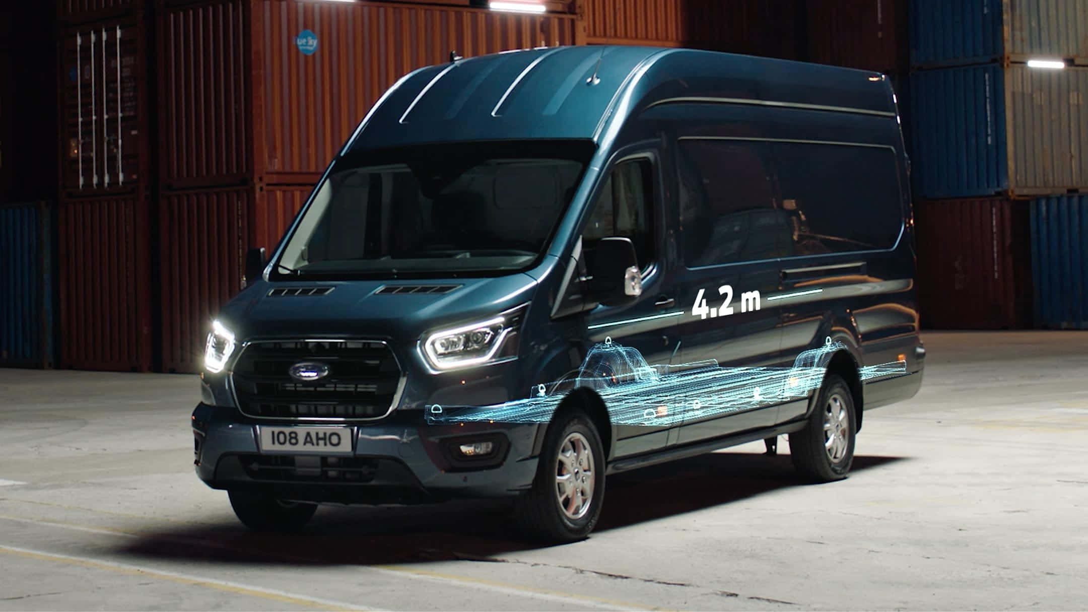 Ford Transit 桌布 壁紙 — 一輛時尚的深藍色福特 Transit 客貨車，車側帶有發光的數位線條和「4.2 m」文字，停放在一個有貨櫃的倉庫中。