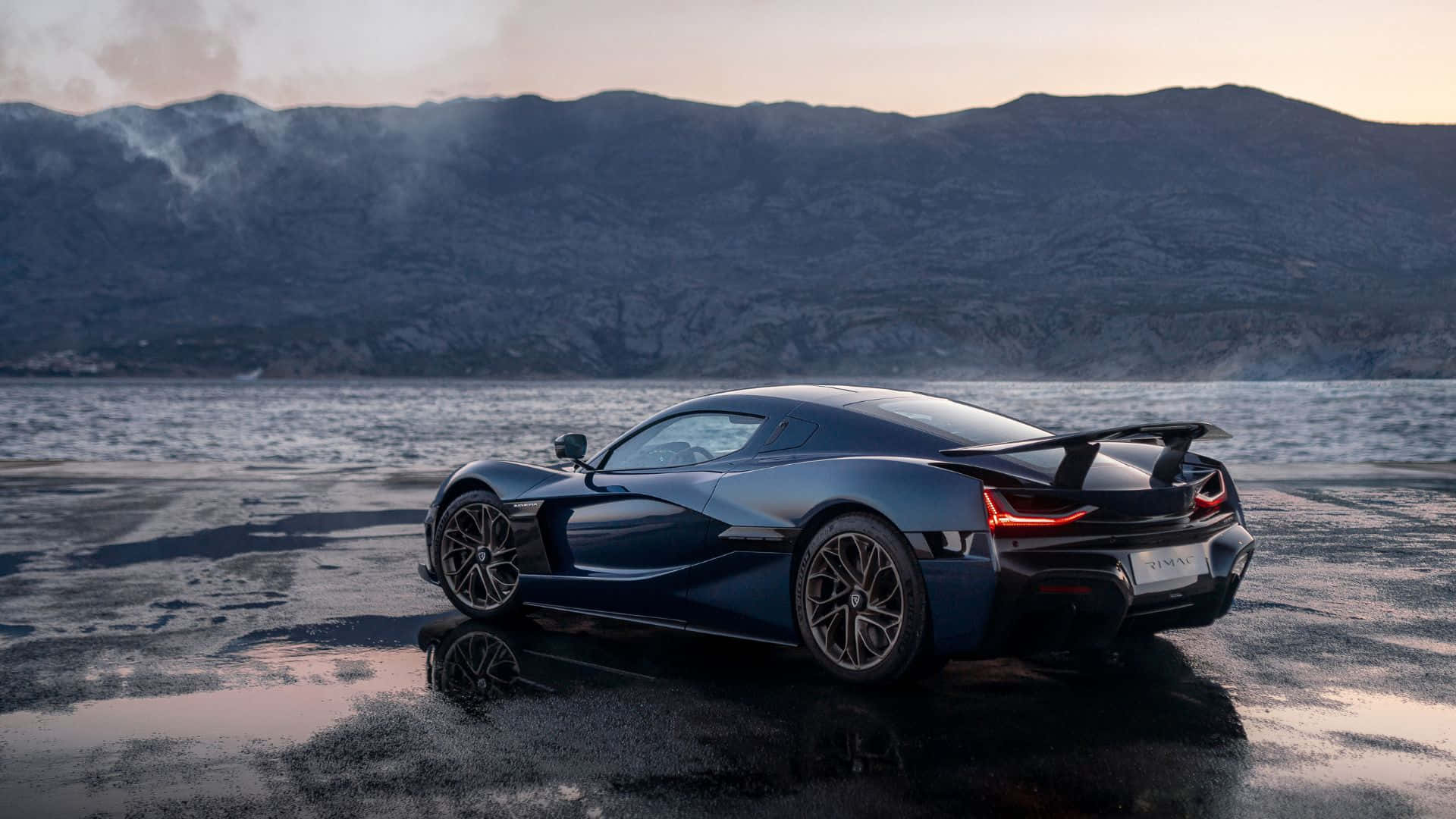 Rimac 桌布 壁紙 — 一輛時尚的深藍色超跑在濕漉漉的路面上反射，旁邊是平靜的大海和霧氣繚繞的山脈，在柔和的暮色天空下。