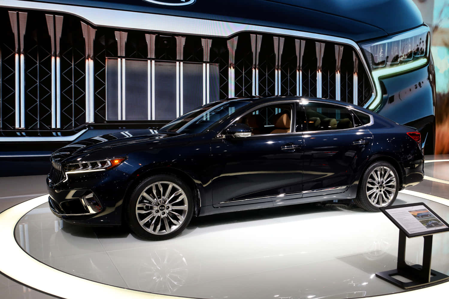 Kia Cadenza 桌布 壁紙 — 一輛時尚的深藍色轎車在圓形展廳舞台上，在明亮燈光下閃閃發光，背景是未來感十足的數位背景。