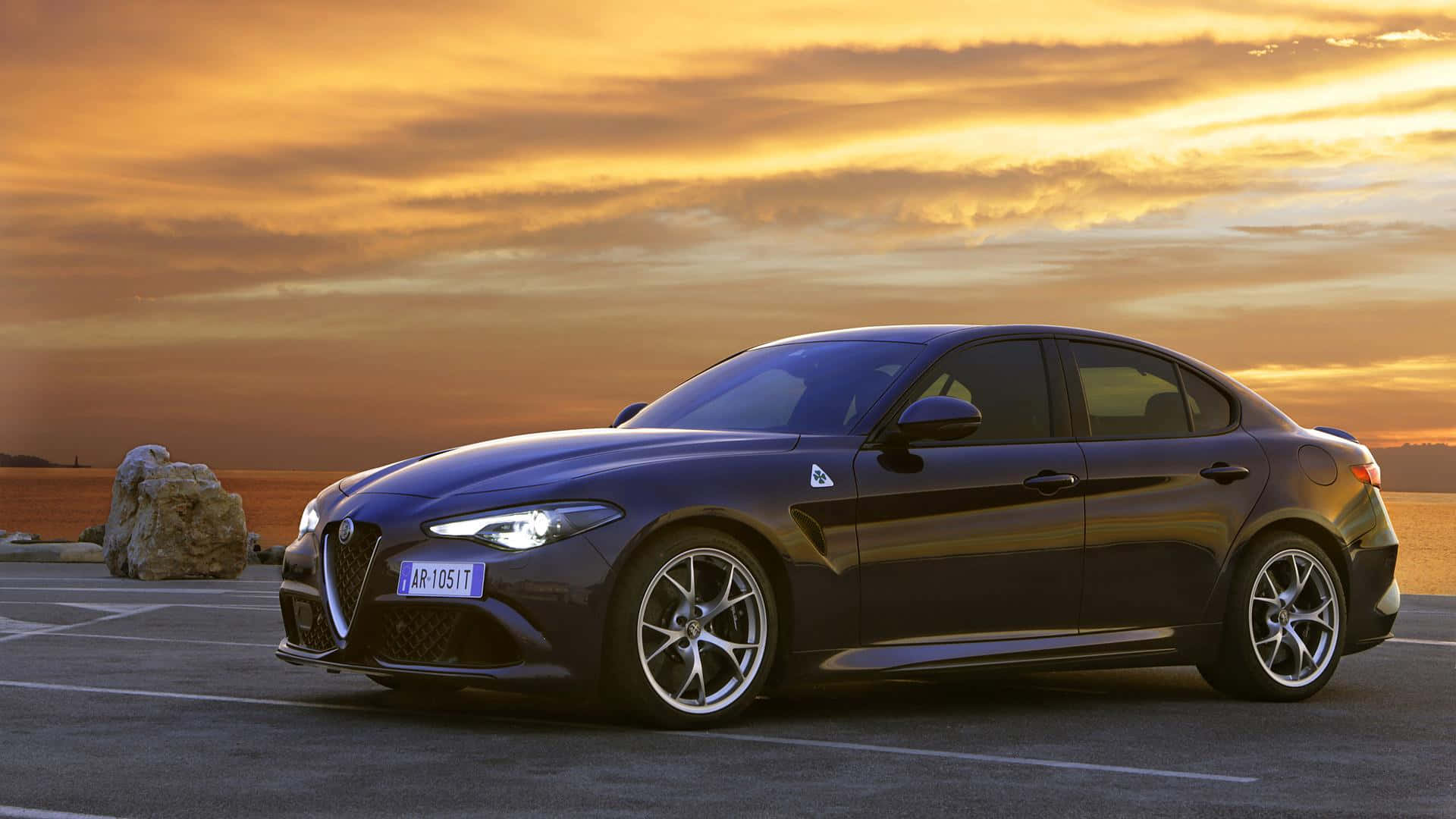 Alfa Romeo Giulia 桌布 壁紙 — 一輛時尚的深藍色阿爾法羅密歐轎車停在日落時分的瀝青路上，附近有金色的雲朵和一塊大岩石。