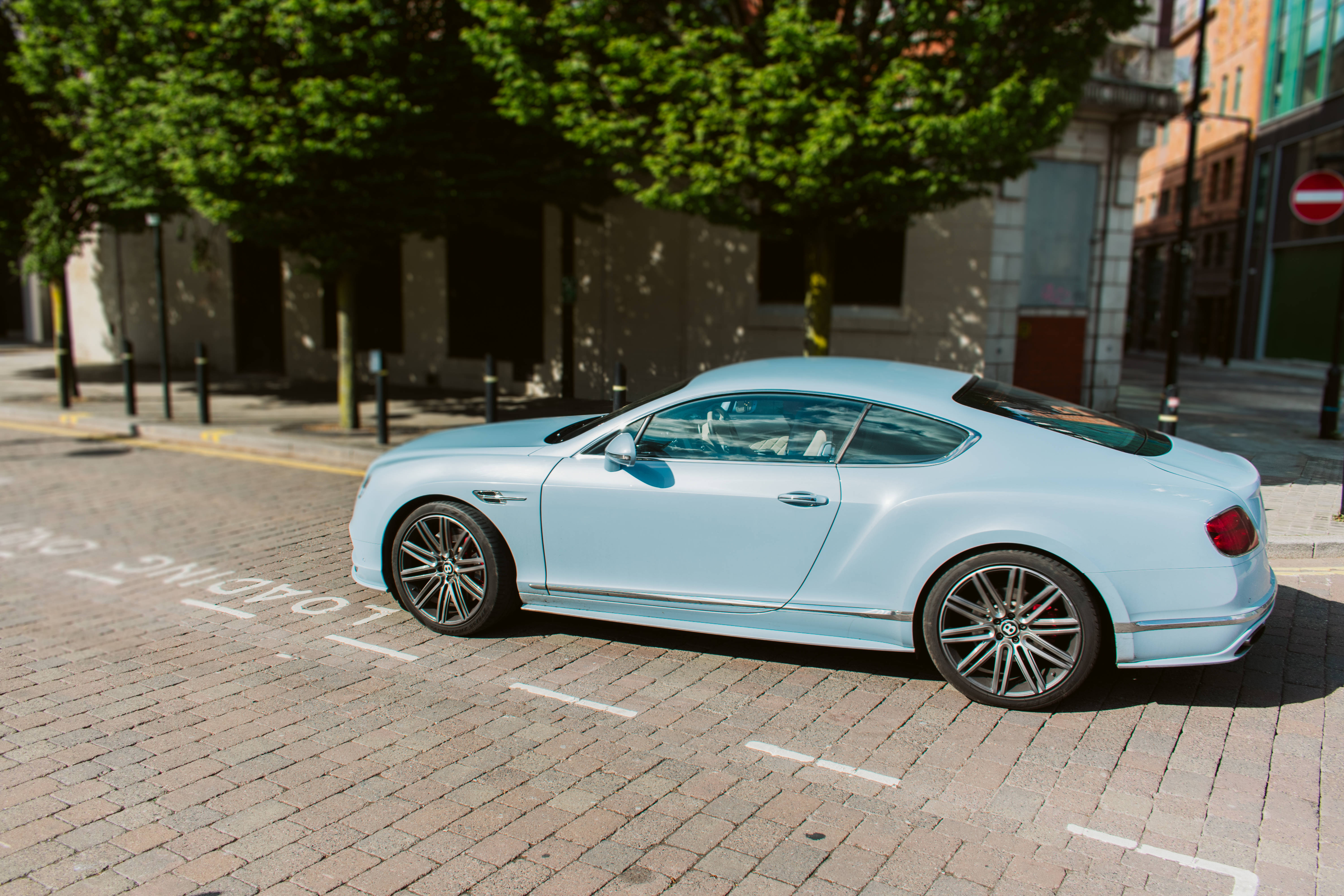 賓利 桌布 壁紙 — 一輛時尚的淺藍色 Bentley Continental GT 停放在陽光明媚的磚砌街道上，兩旁有綠樹和城市建築。
