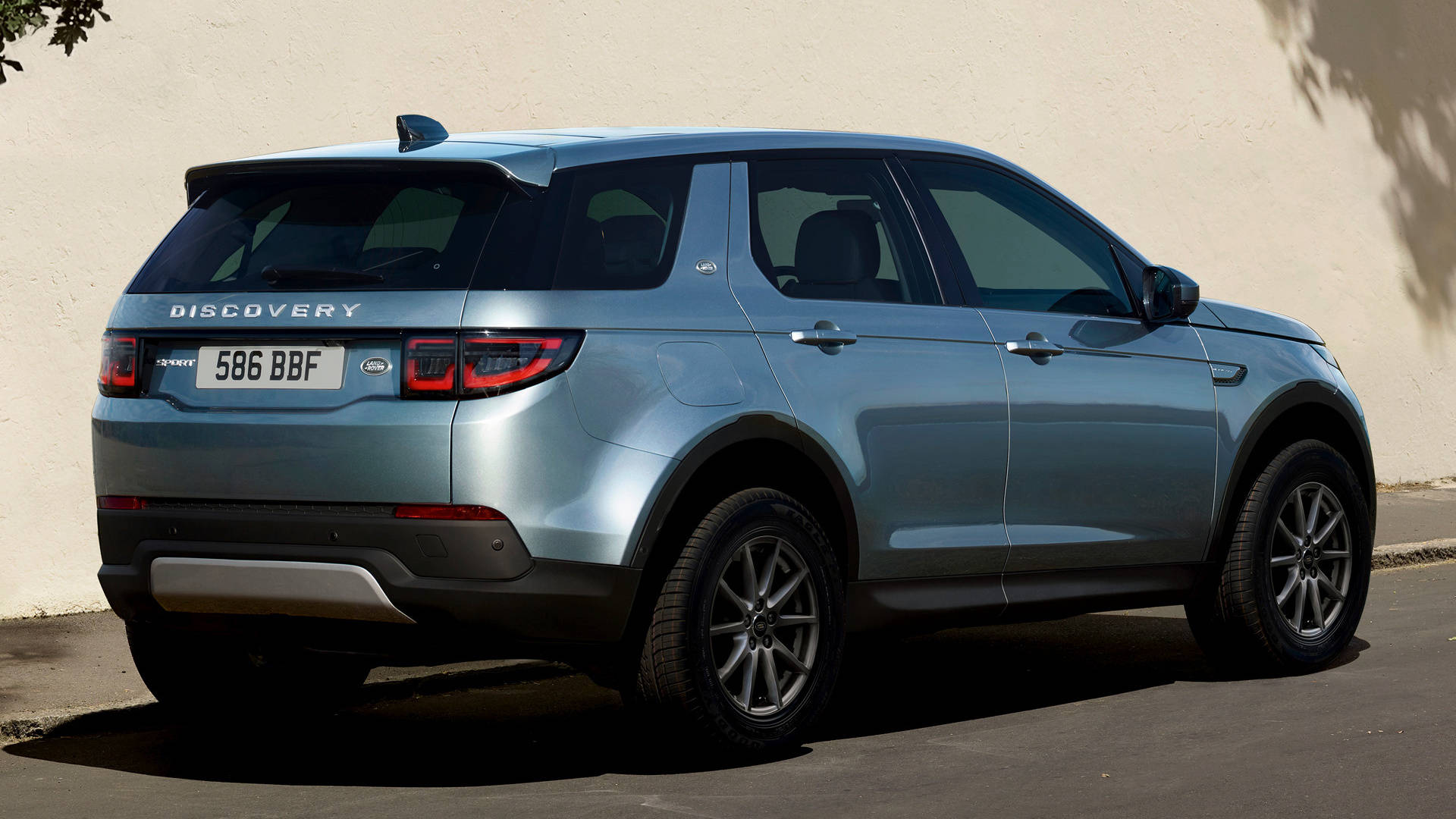 land rover iPhone 桌布 壁紙 — 一輛時尚的淺藍色 Land Rover Discovery SUV 以傾斜角度停放在陽光照射的米色牆壁旁，投射出銳利的陰影。