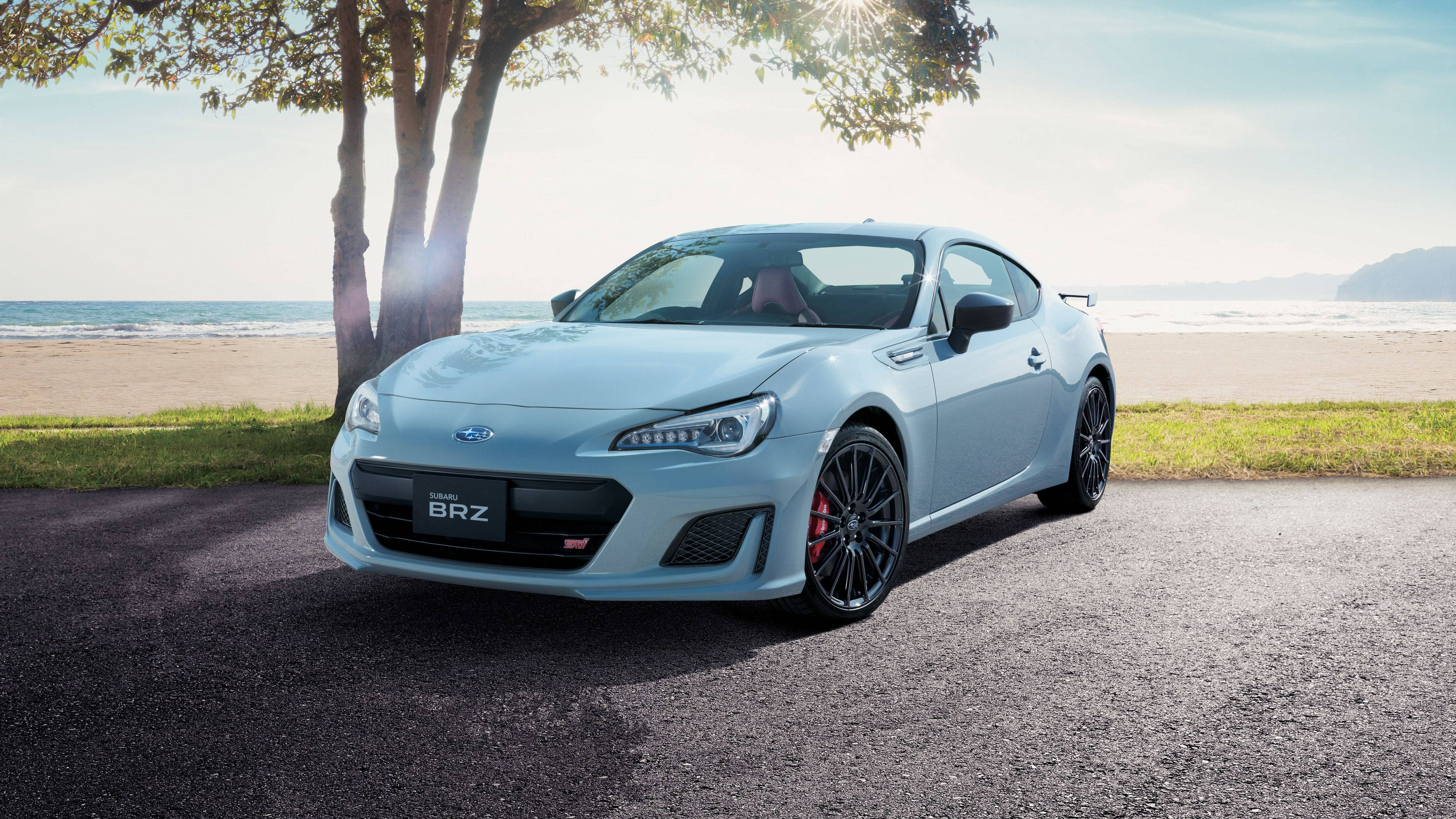昴 桌布 壁紙 — 一輛時尚的淺藍色Subaru BRZ跑車停放在鋪砌小徑上，旁邊有樹木，背景是海洋和沙灘，在明亮的陽光下。
