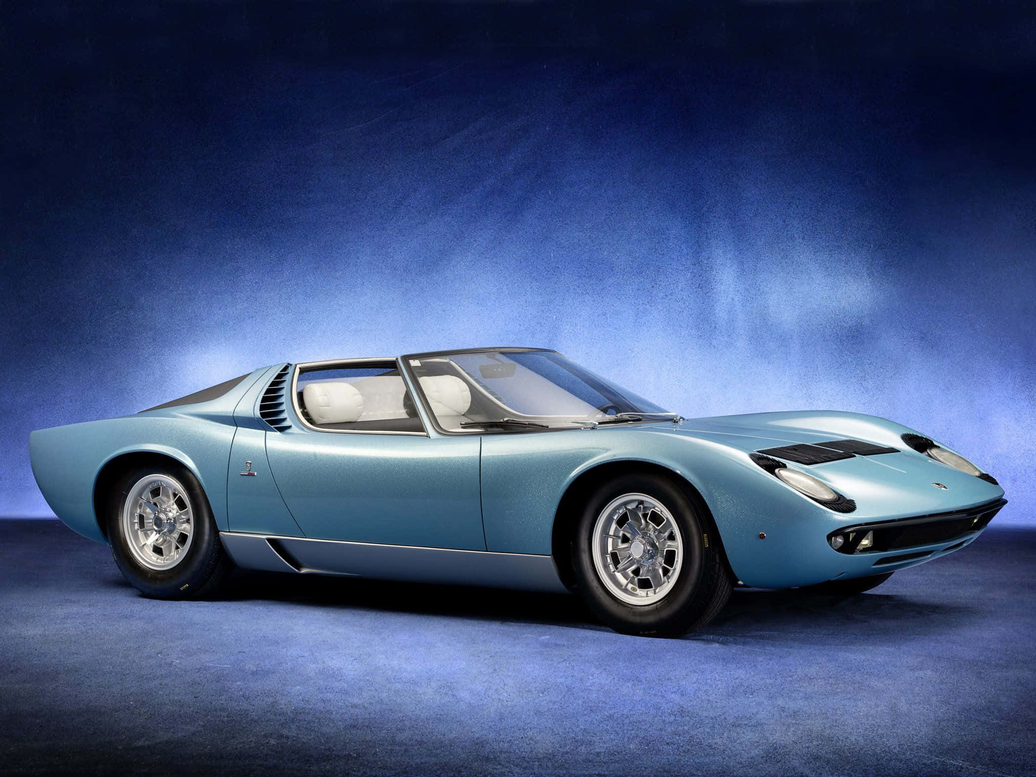 Lamborghini Miura 桌布 壁紙 — 一輛時尚的淺藍色復古跑車，帶有銀色飾邊和白色內飾，在戲劇性的藍色漸變工作室背景前擺拍。