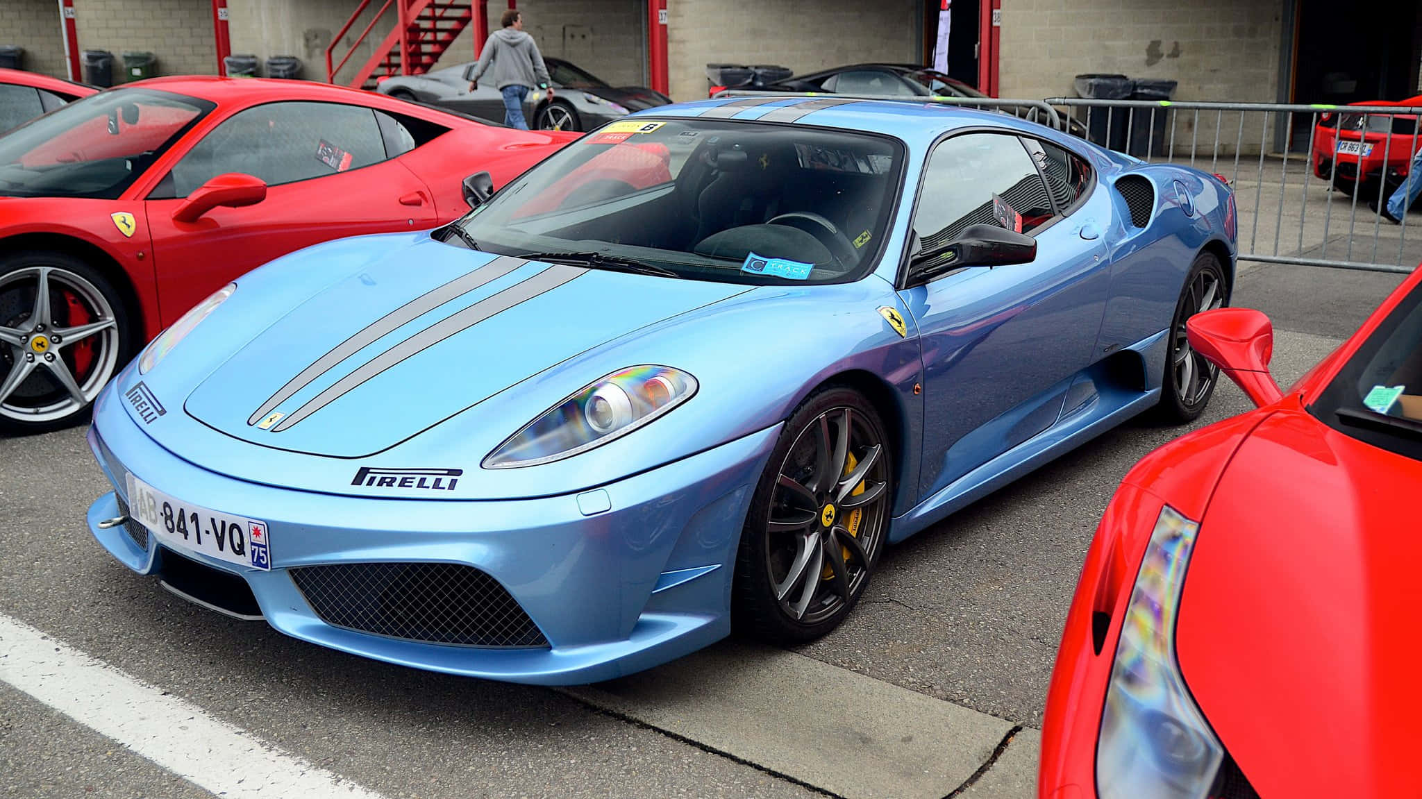 Ferrari F430 桌布 壁紙 — 一輛時尚的淺藍色法拉利跑車，帶有灰色賽車條紋，停放在車庫中的紅色法拉利之間。