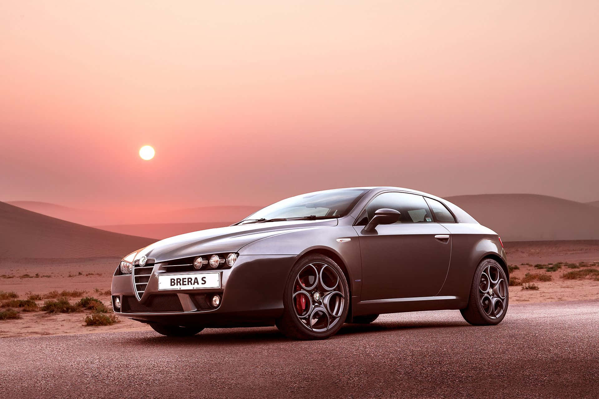 Alfa Romeo Brera 桌布 壁紙 — 一輛時尚的灰色 Alfa Romeo Brera S 跑車停泊在沙漠公路上，夕陽西下，橙色的太陽在連綿沙丘後方閃耀。