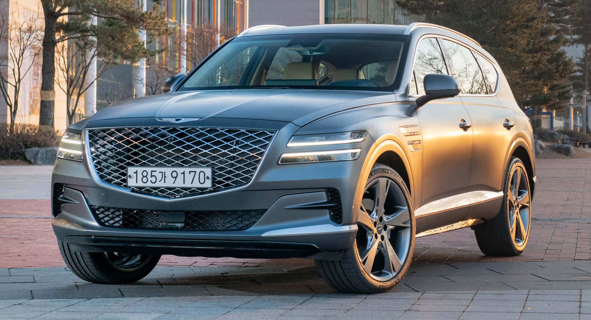 Genesis Gv80 桌布 壁紙 — 一輛時尚的灰色 Genesis GV80 SUV 停放在鋪砌的廣場上，周圍有樹木和建築物，沐浴在柔和的晨光中。