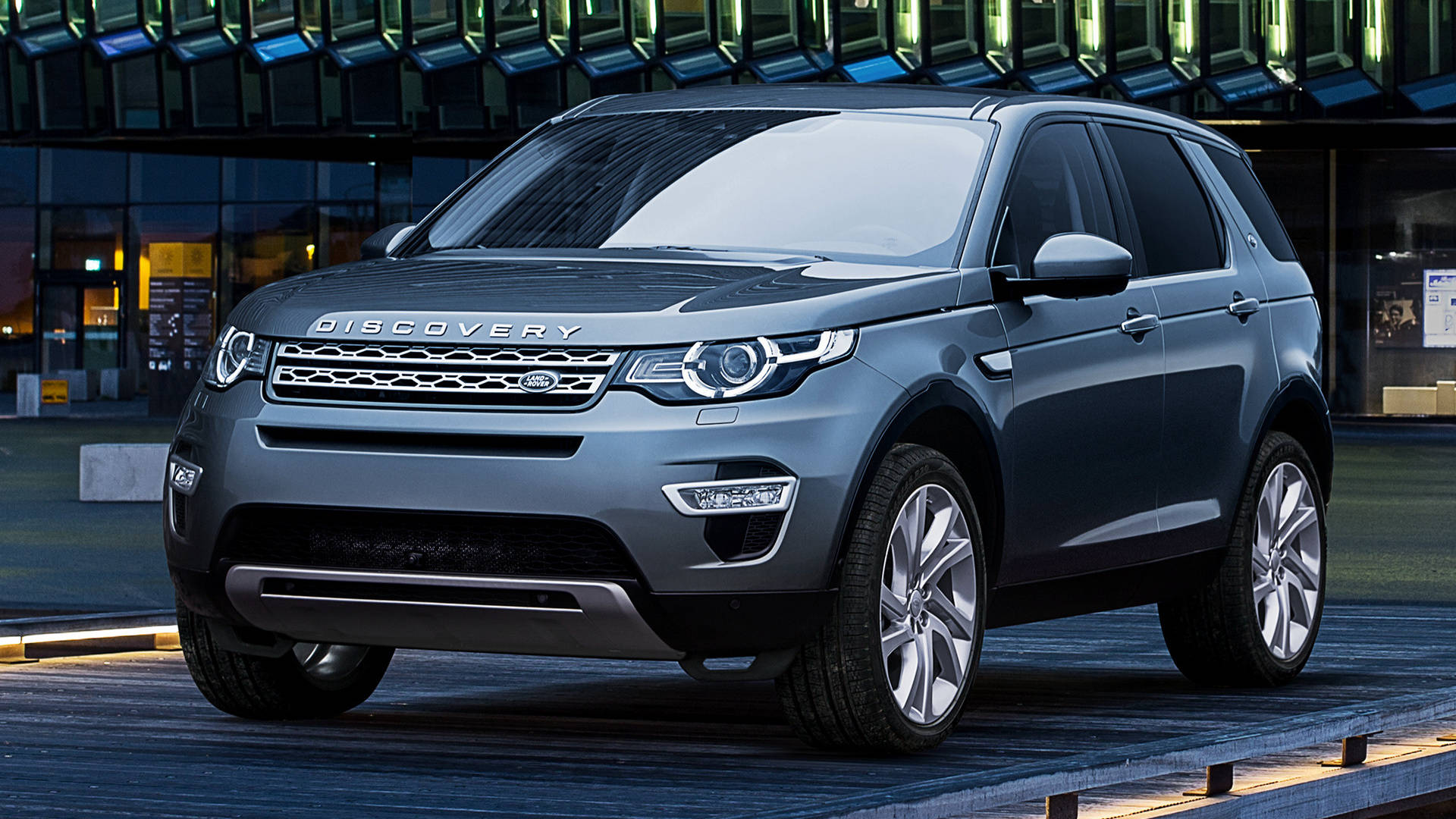 land rover iPhone 桌布 壁紙 — 一輛時尚的灰色 Land Rover Discovery SUV 停泊在黃昏時分現代玻璃建築前的木質平台上。