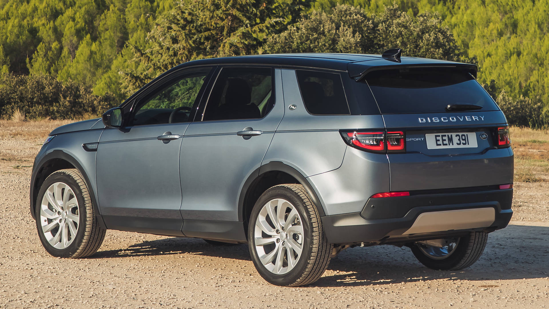 land rover iPhone 桌布 壁紙 — 一輛時尚的灰色 Land Rover Discovery Sport 停在碎石路上，背景是翠綠的樹木，在明亮的陽光下。