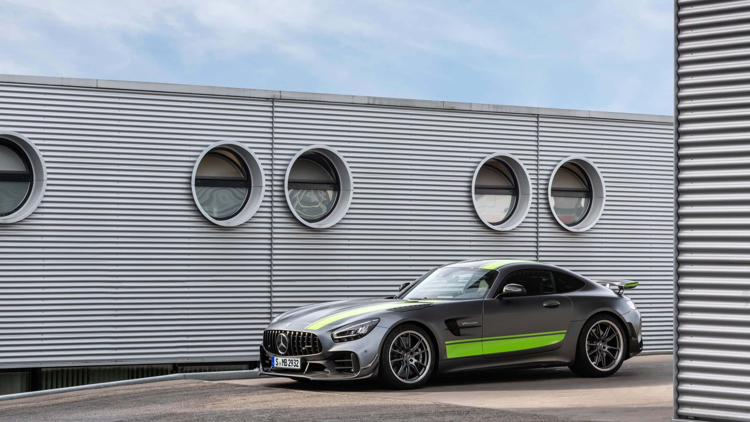 Mercedes 桌布 壁紙 — 一輛時尚的灰色 Mercedes-AMG GT 帶有螢光綠色點綴，停在一個帶有圓形窗戶的現代波紋金屬建築前。