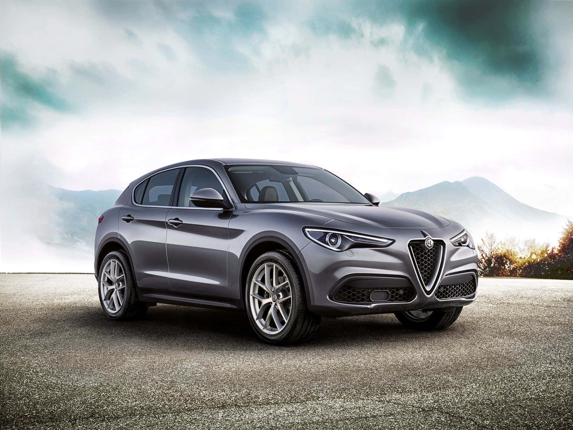 Alfa Romeo Stelvio 桌布 壁紙 — 一輛時尚的灰色Alfa Romeo SUV在戲劇性的多雲天空下，停泊在霧氣瀰漫的山路上。