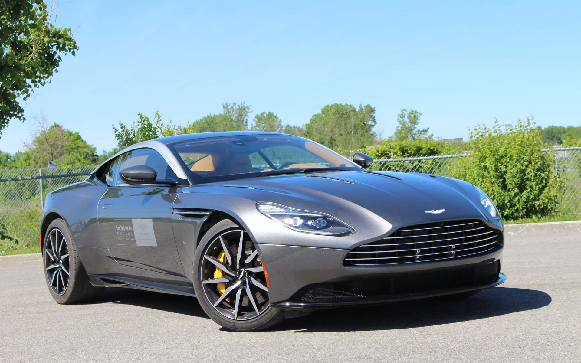 奧斯頓馬丁 桌布 壁紙 — 一輛時尚的灰色Aston Martin跑車停在柏油路上，周圍有綠樹和鐵絲網圍欄，天空湛藍。