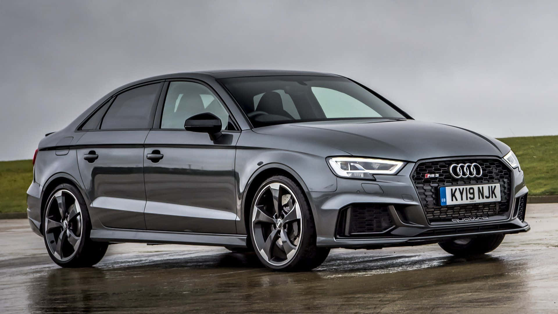 Sedan 桌布 壁紙 — 一輛時尚的灰色Audi RS3轎車搭配黑色輪圈，停在濕滑的道路上，背景是陰天和長滿草的山丘。