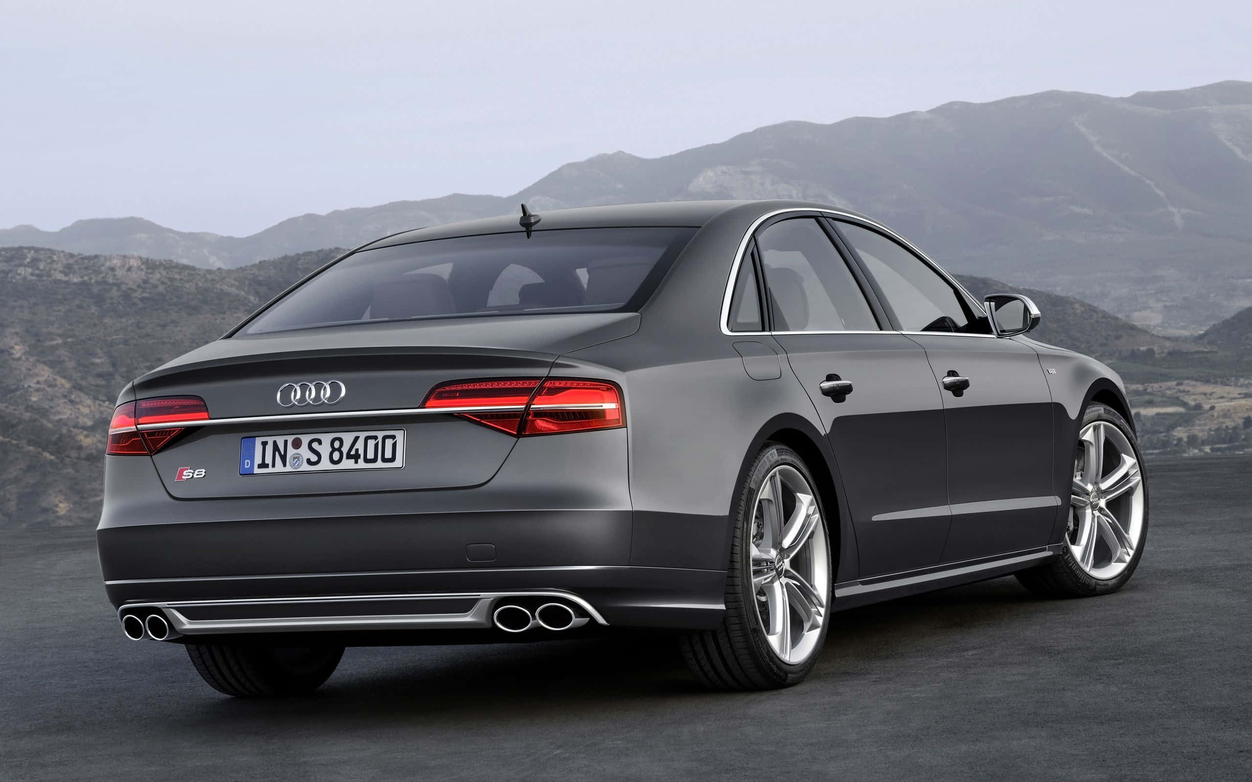 Audi S8 桌布 壁紙 — 一輛時尚的灰色Audi S8轎車停放在鋪砌的道路上，背景是崎嶇的山脈，天空朦朧。