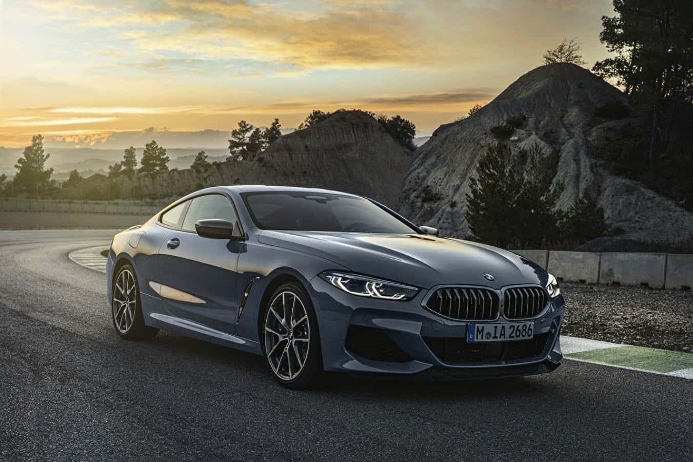 跑車 桌布 壁紙 — 一輛時尚的灰色BMW 8系列轎跑車在金色日落、戲劇性的天空和岩石地形中沿著蜿蜒的山路滑行。
