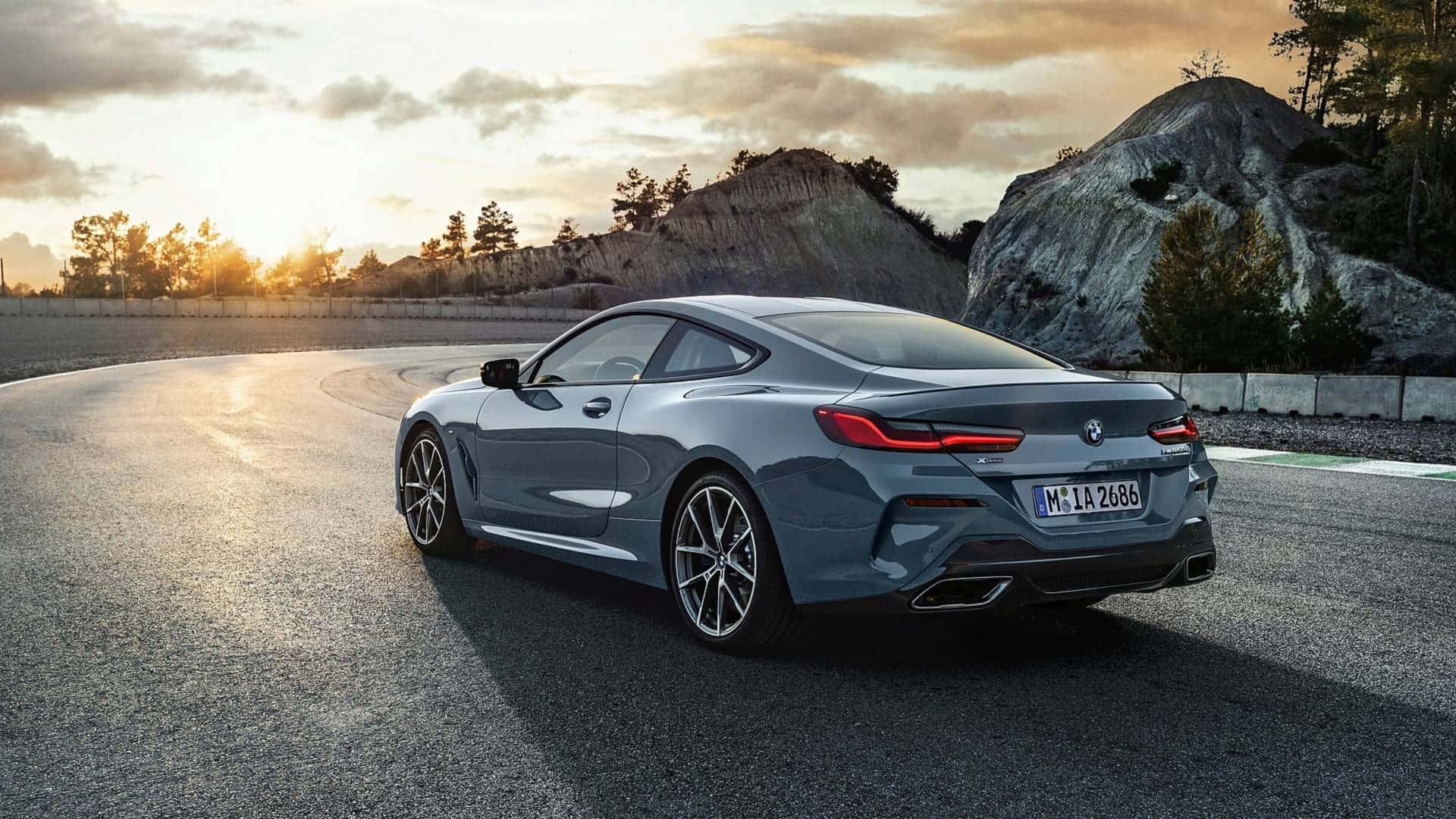 Bmw 8 Series 桌布 壁紙 — 一輛時尚的灰色BMW 8系列轎跑車在日落時分沿著蜿蜒的瀝青道路行駛，背景是岩石丘陵和樹木。