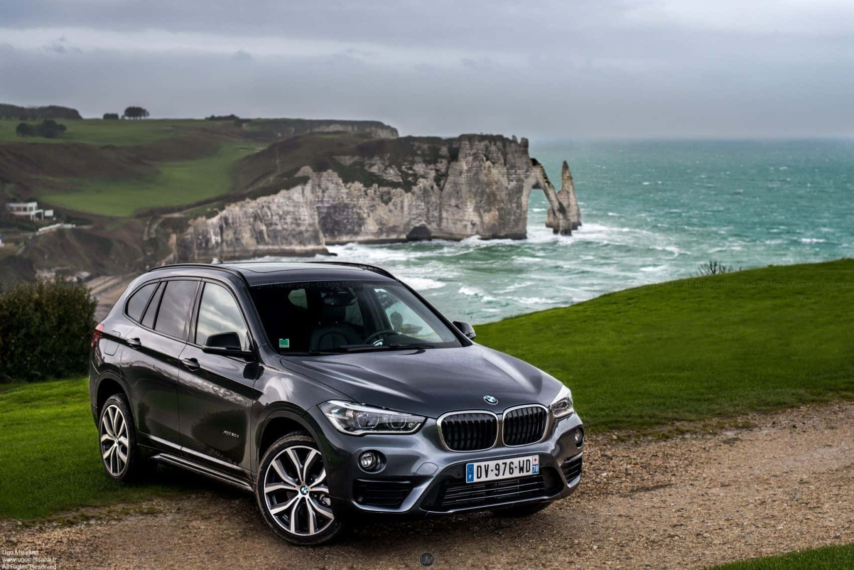 Bmw X1 桌布 壁紙 — 一輛時尚的灰色BMW SUV停泊在俯瞰海洋的草地上懸崖上，背景是標誌性的白色白堊懸崖和拱門，天空多雲。