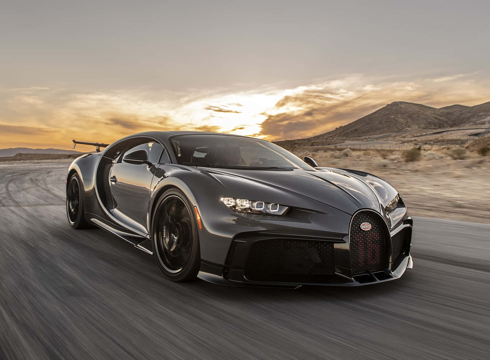 Best Bugatti 桌布 壁紙 — 一輛時尚的灰色Bugatti Chiron在沙漠賽道上於日落時分疾馳，伴隨著戲劇性的動態模糊和金色天空。