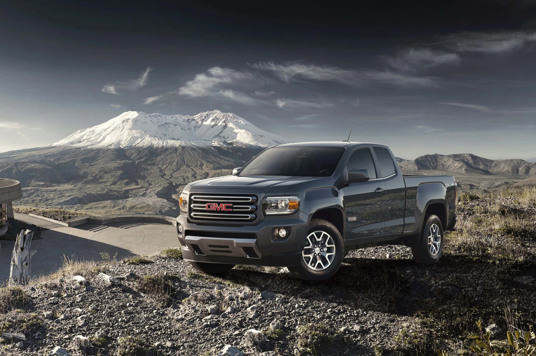 Gmc Canyon 桌布 壁紙 — 一輛時尚的灰色GMC Sierra皮卡停泊在岩石觀景點上，背後有白雪覆蓋的山峰在戲劇性的多雲天空下升起。