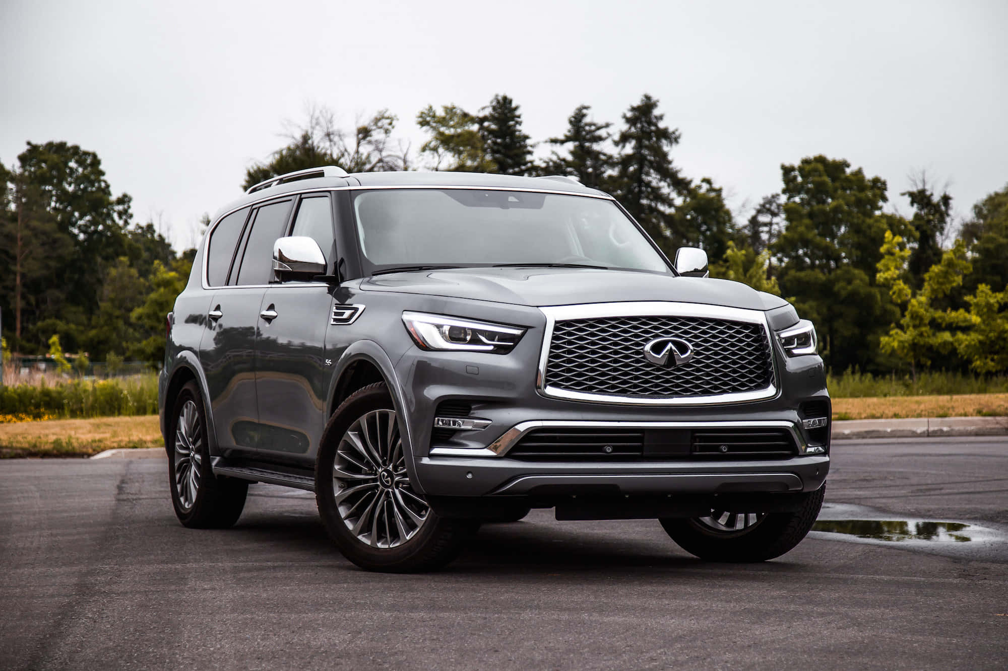 Infiniti Qx80 桌布 壁紙 — 一輛時尚的灰色Infiniti QX80 SUV停在潮濕的路面上，背景有樹木，天空多雲。