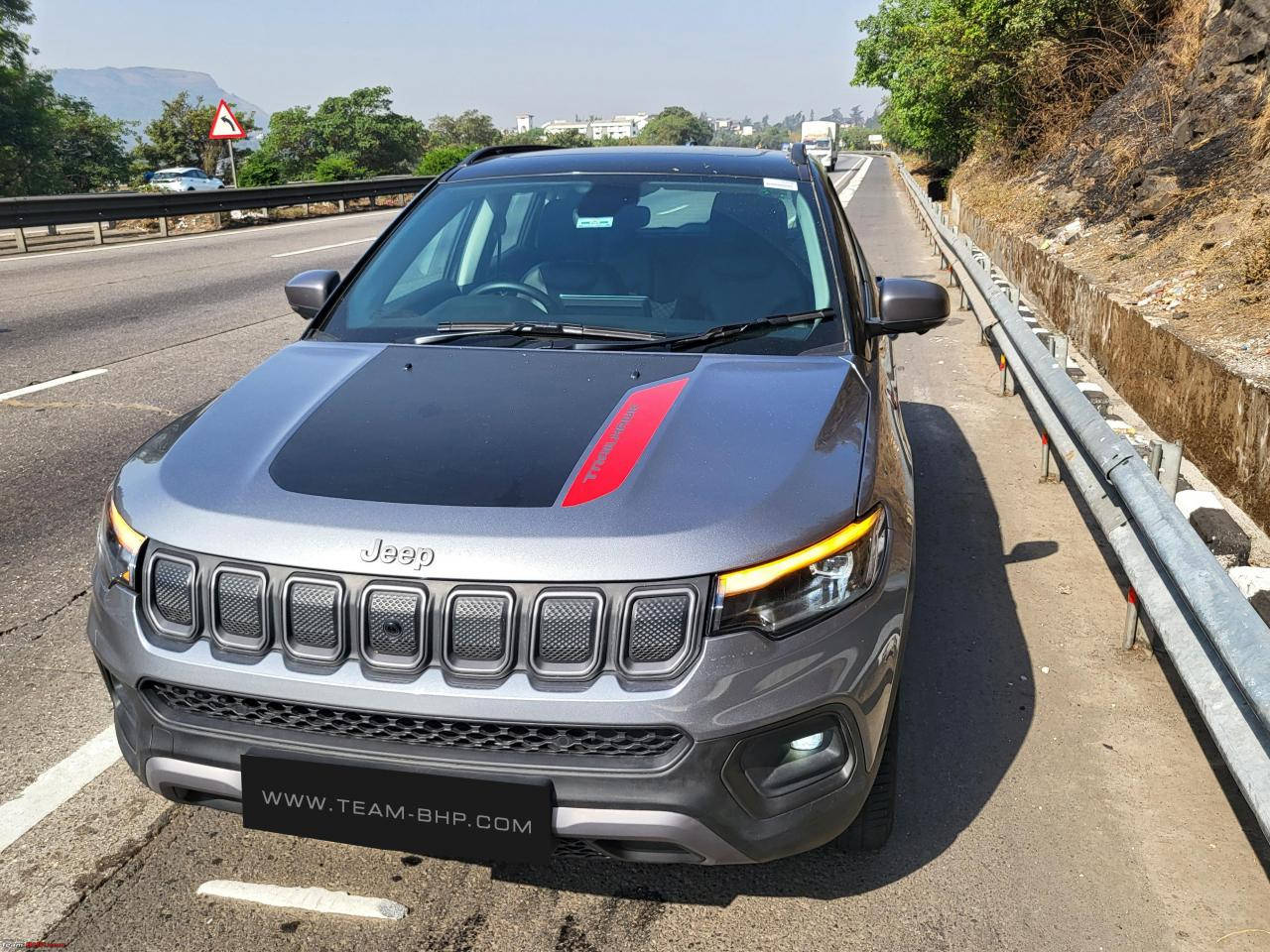 Road 桌布 壁紙 — 一輛時尚的灰色Jeep Compass，引擎蓋上有紅色條紋，停在陽光明媚的高速公路旁，旁邊有綠樹和遠山。