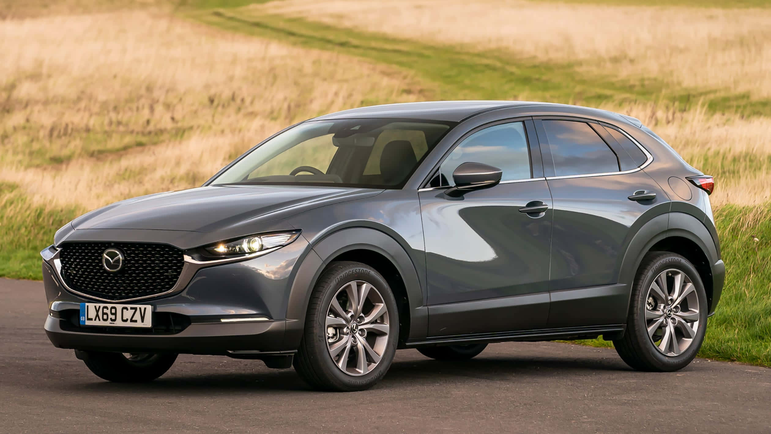 Mazda Cx-30 桌布 壁紙 — 一輛時尚的灰色Mazda CX-3停在路邊，旁邊是金黃色的草地，在明亮的日光下。