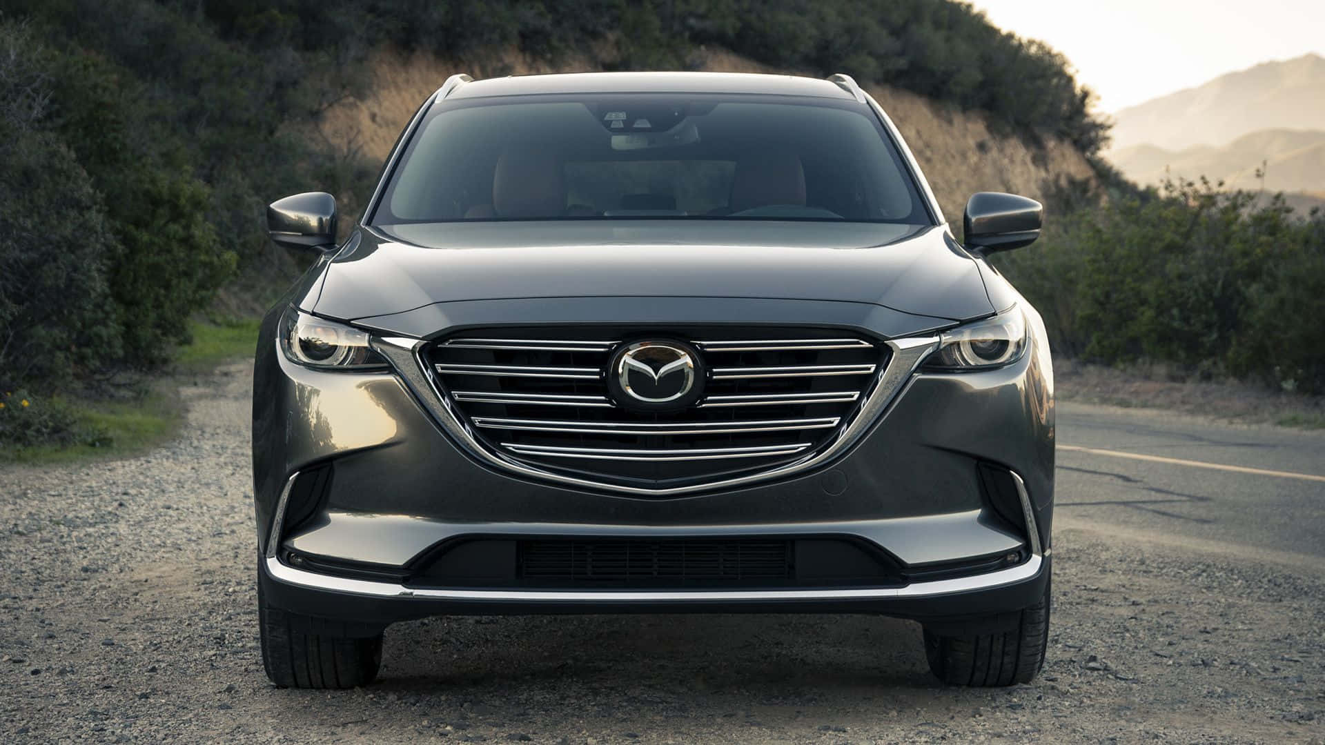 Mazda Cx-9 桌布 壁紙 — 一輛時尚的灰色Mazda CX-9 SUV在碎石路上向前行駛，背景是丘陵和樹木，沐浴在柔和的金色光線下。