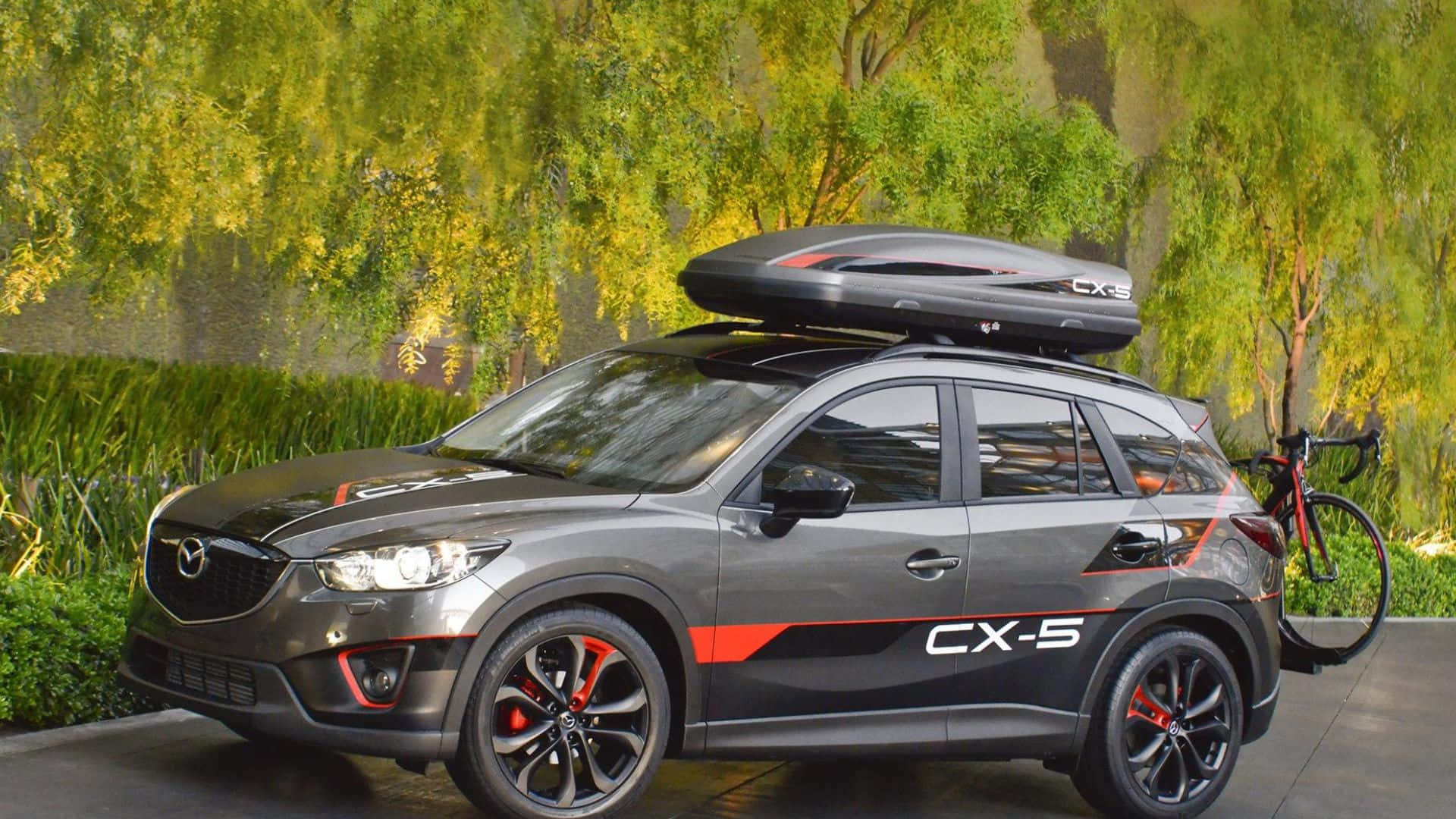 Mazda Cx-5 桌布 壁紙 — 一輛時尚的灰色Mazda CX-5，帶有紅色點綴和車頂箱，停在鮮綠色垂柳樹旁的紅色自行車旁邊。