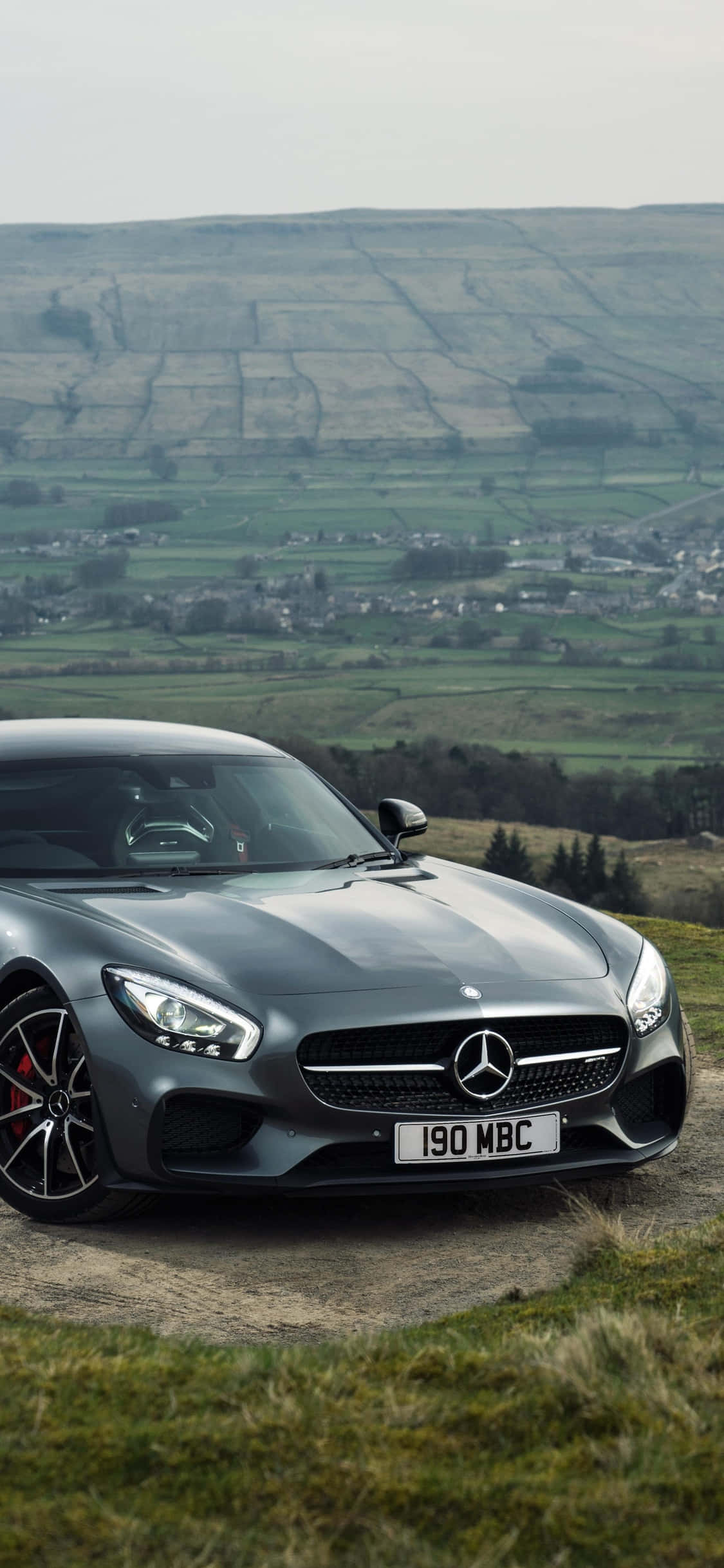 iPhone X Amg Gt-r 桌布 壁紙 — 一輛時尚的灰色Mercedes-AMG GT停泊在一個草坡上，俯瞰著一個廣闊的鄉村山谷，山谷中有拼布般的田野和遠處的村莊，天空多雲。