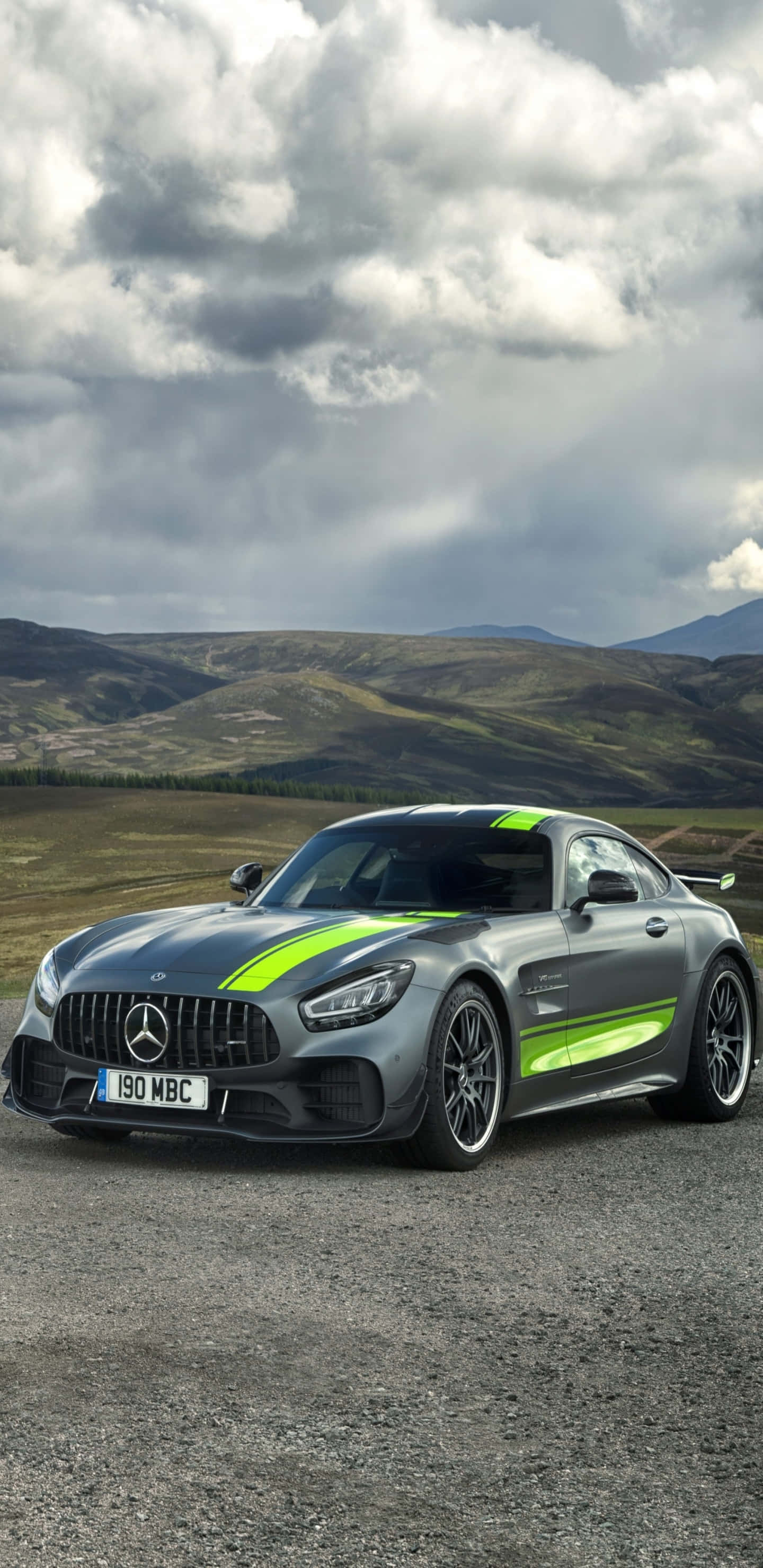 Black Car 桌布 壁紙 — 一輛時尚的灰色Mercedes-AMG GT R，帶有螢光綠色條紋，停在碎石上，背景是戲劇性的多雲天空和起伏的山丘。