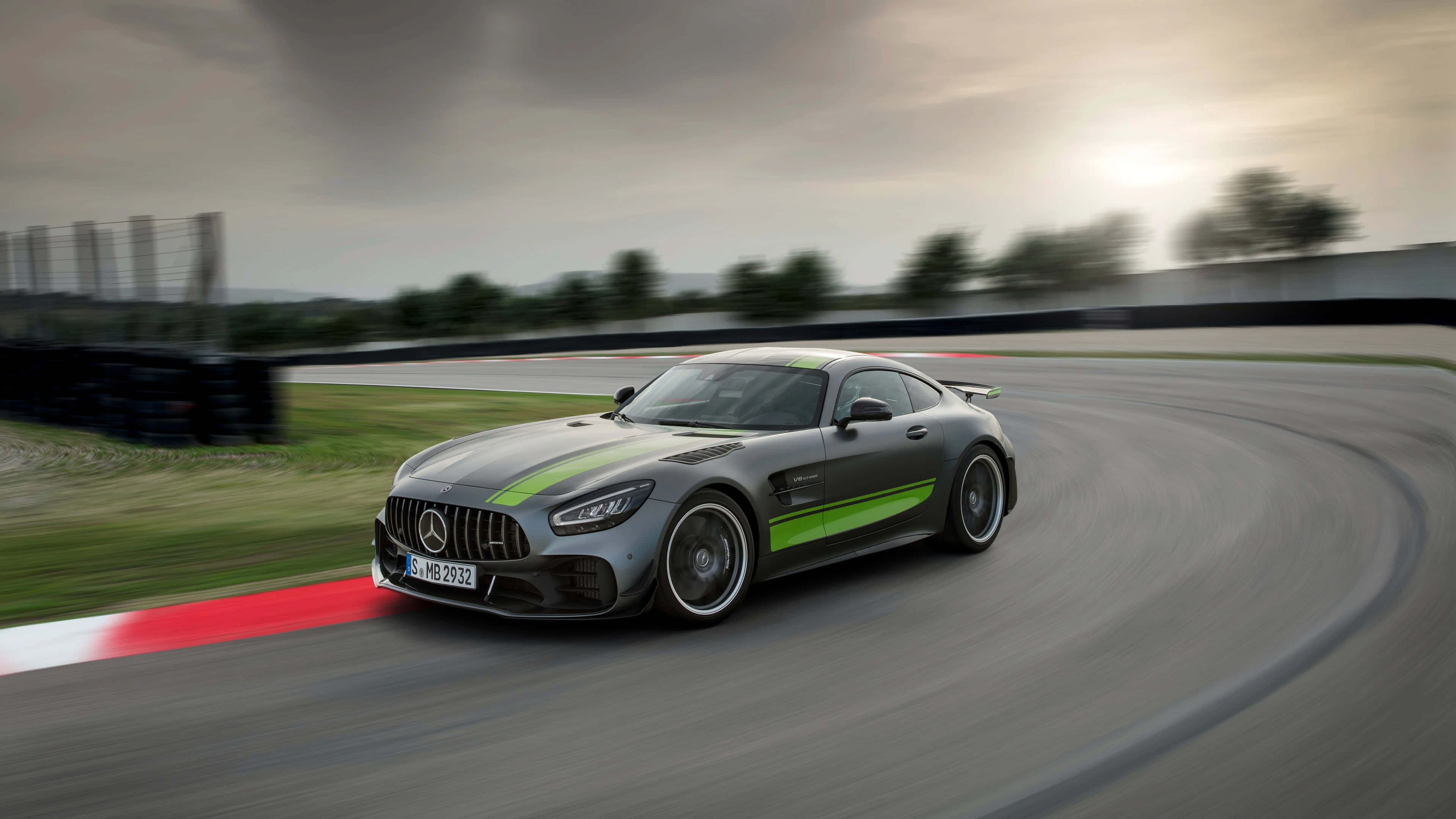 Mercedes 桌布 壁紙 — 一輛時尚的灰色Mercedes-AMG GT賽車，帶有綠色條紋，在多雲的天空下沿著彎曲的賽道飛馳。