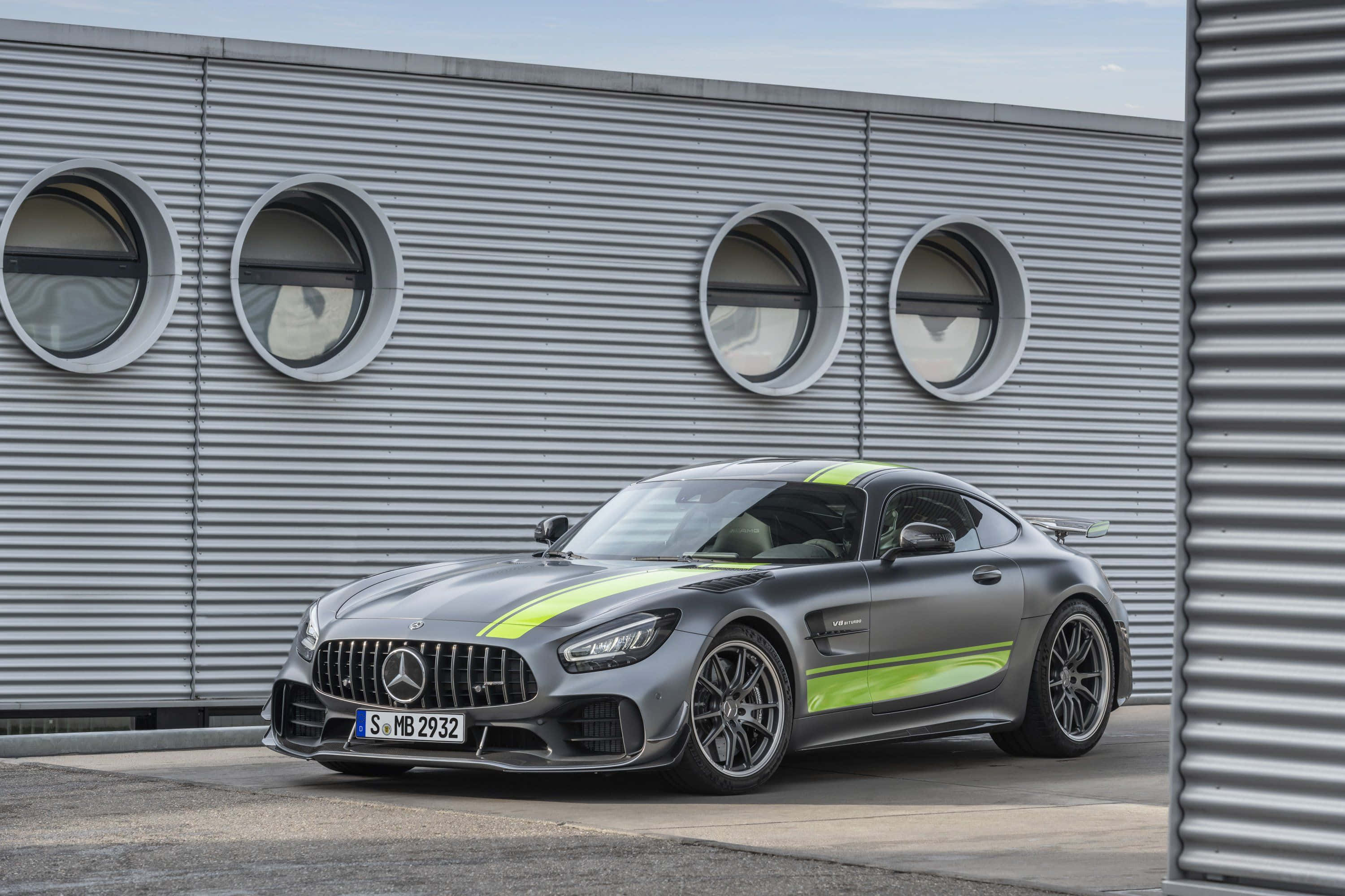 跑車 桌布 壁紙 — 一輛時尚的灰色Mercedes-AMG GT，帶有螢光綠色條紋，停在帶有圓形窗戶的波紋金屬牆前。
