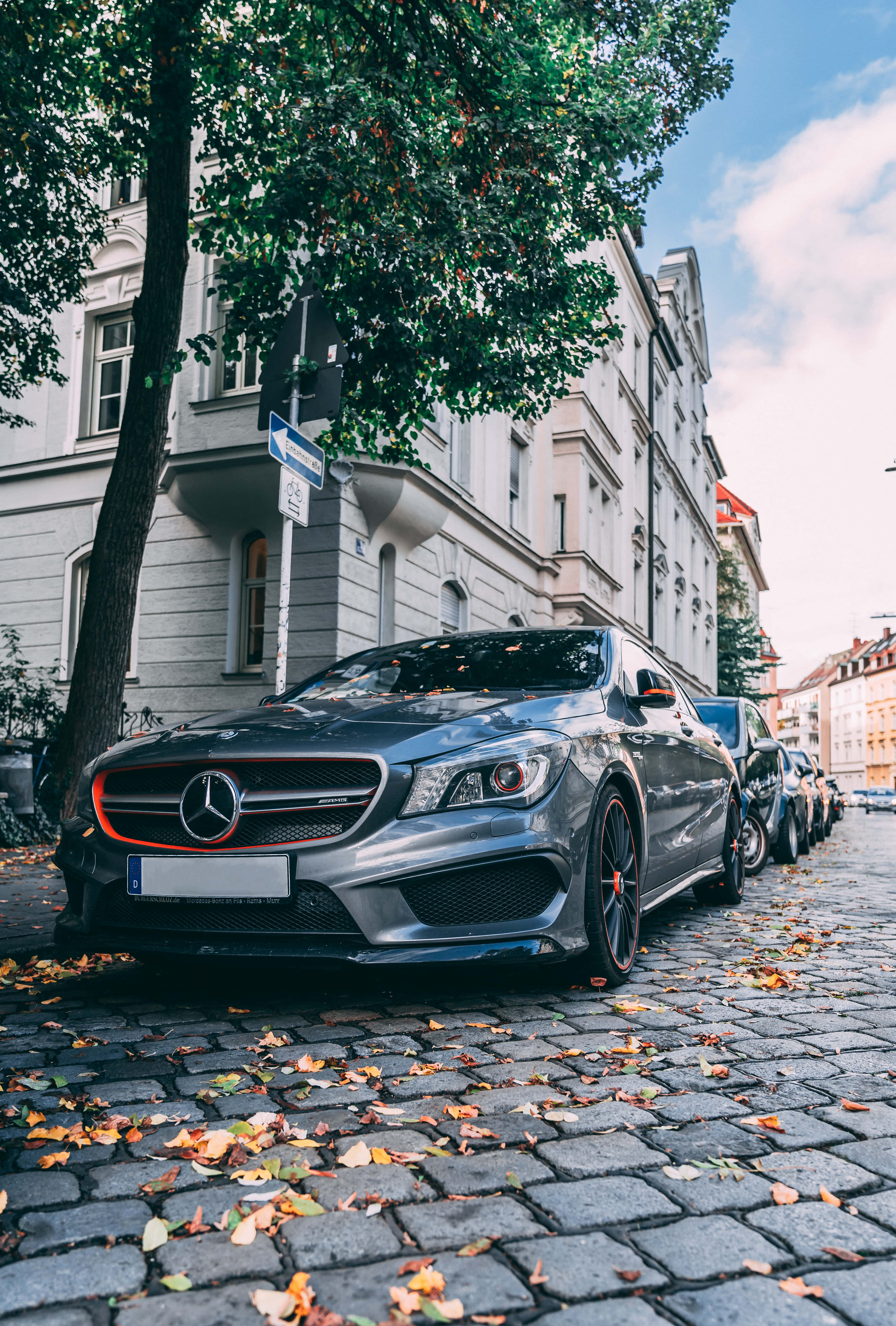 iPhone X Mercedes 桌布 壁紙 — 一輛時尚的灰色Mercedes-AMG CLA45停泊在散落著秋葉的鵝卵石街道上，天空部分多雲。