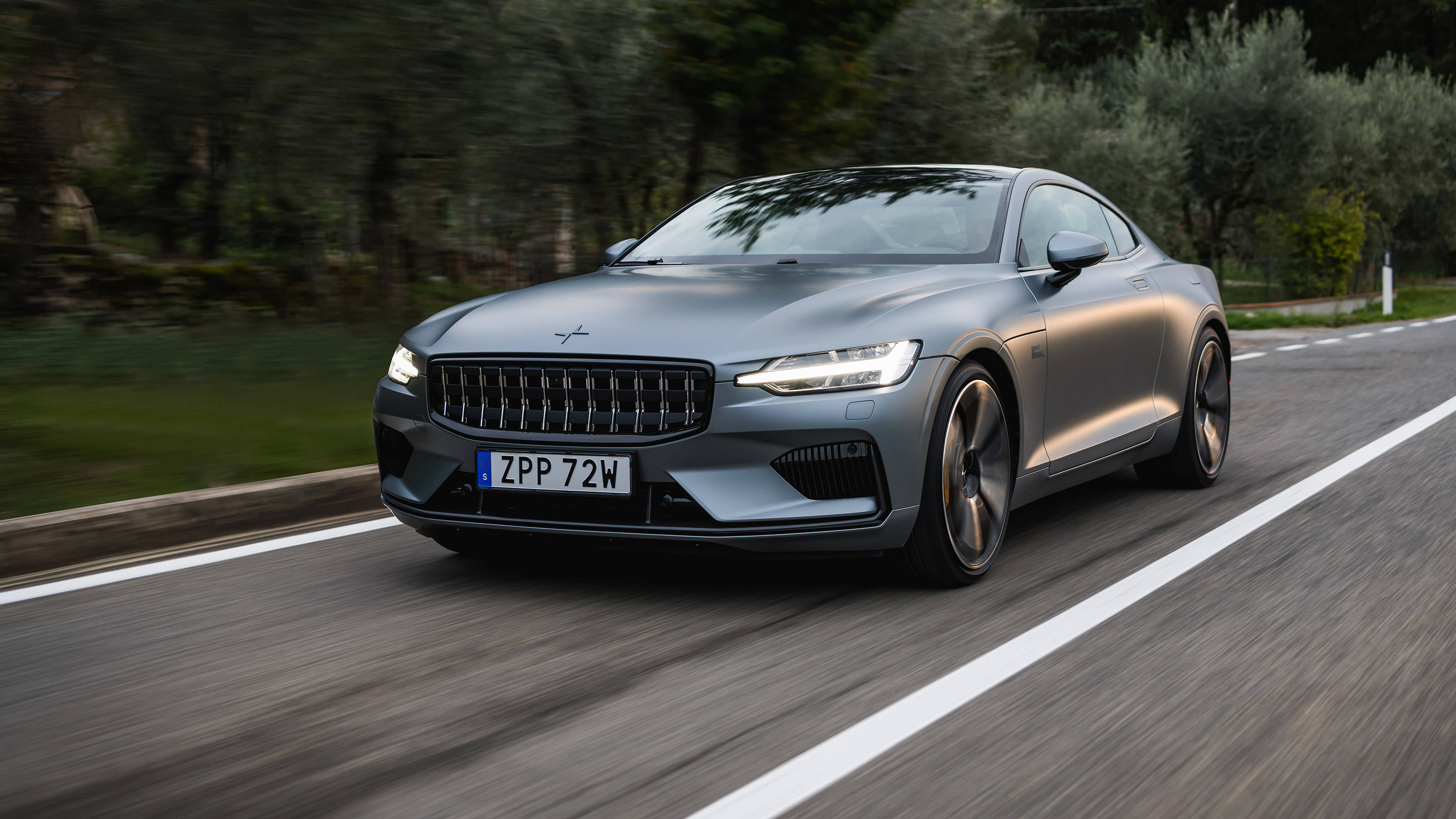 豪華車 桌布 壁紙 — 一輛時尚的灰色Polestar 2跑車沿著蜿蜒的道路穿過茂密的綠色森林飛馳。