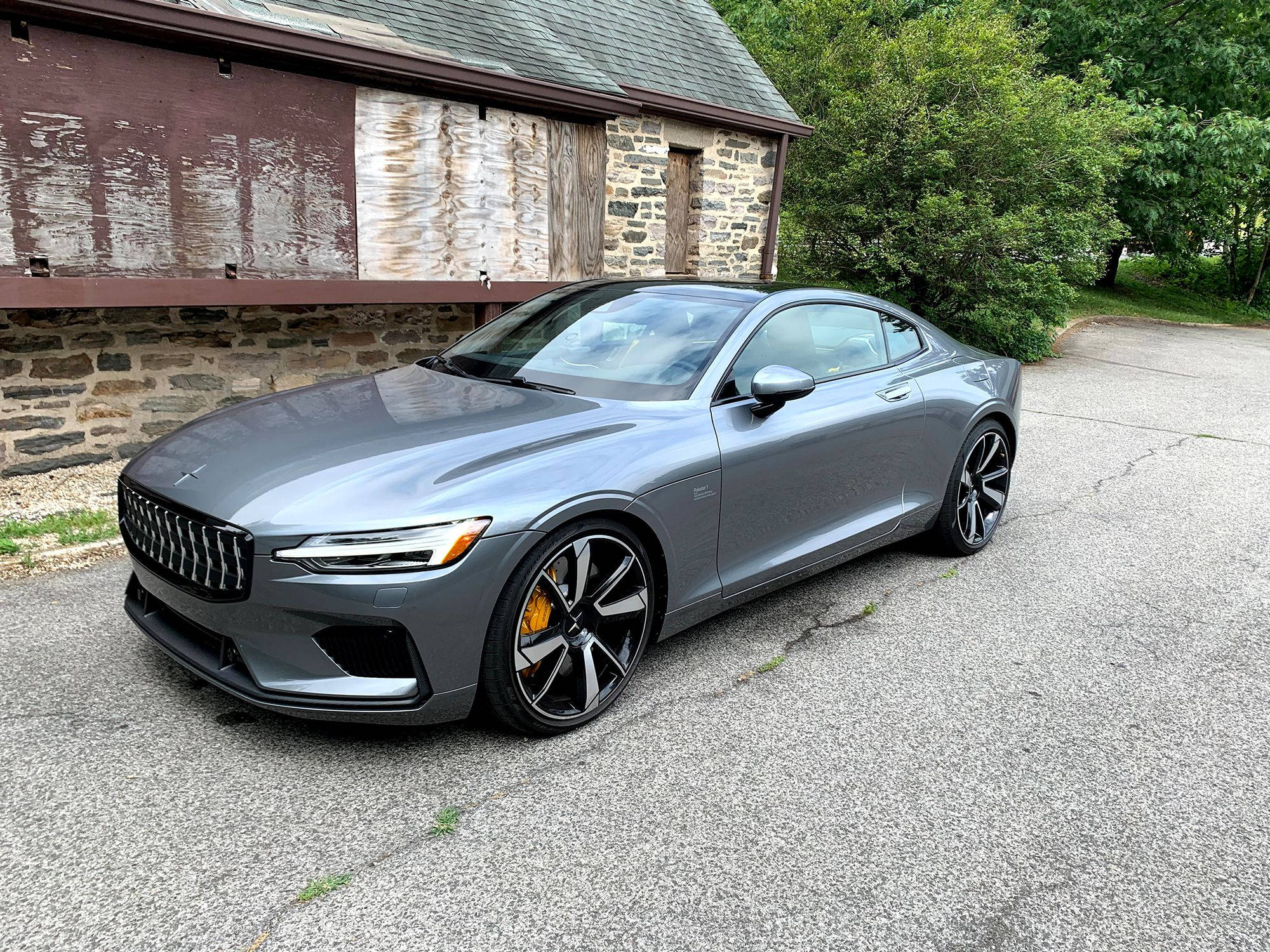 豪華車 桌布 壁紙 — 一輛時尚的灰色Polestar 2電動轎跑車停在裂開的瀝青車道上，旁邊是一座質樸的石木建築，背景是茂密的綠樹。