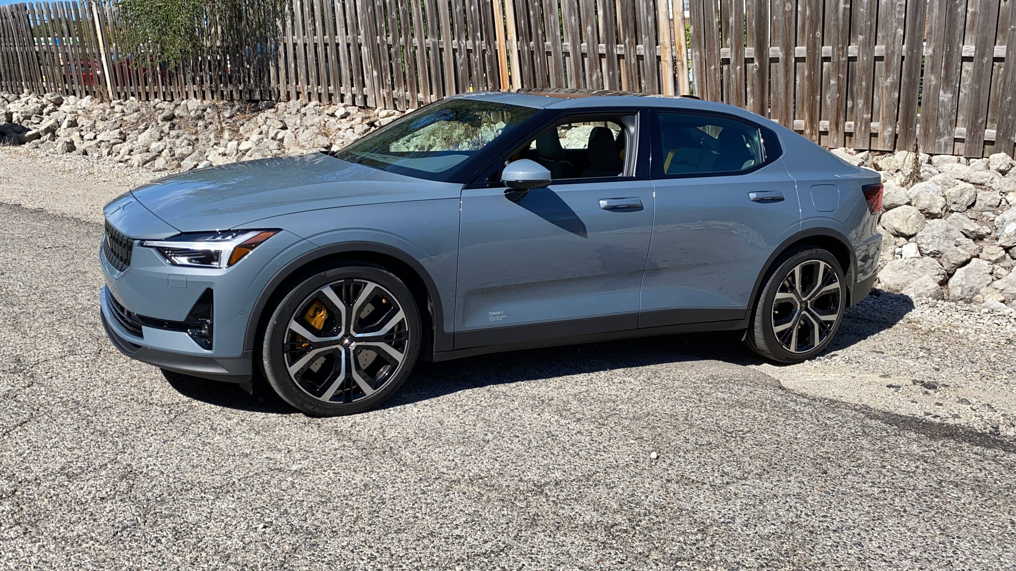 豪華車 桌布 壁紙 — 一輛時尚的灰色Polestar 2電動車停在碎石上，旁邊是木柵欄和石牆，陽光明媚。