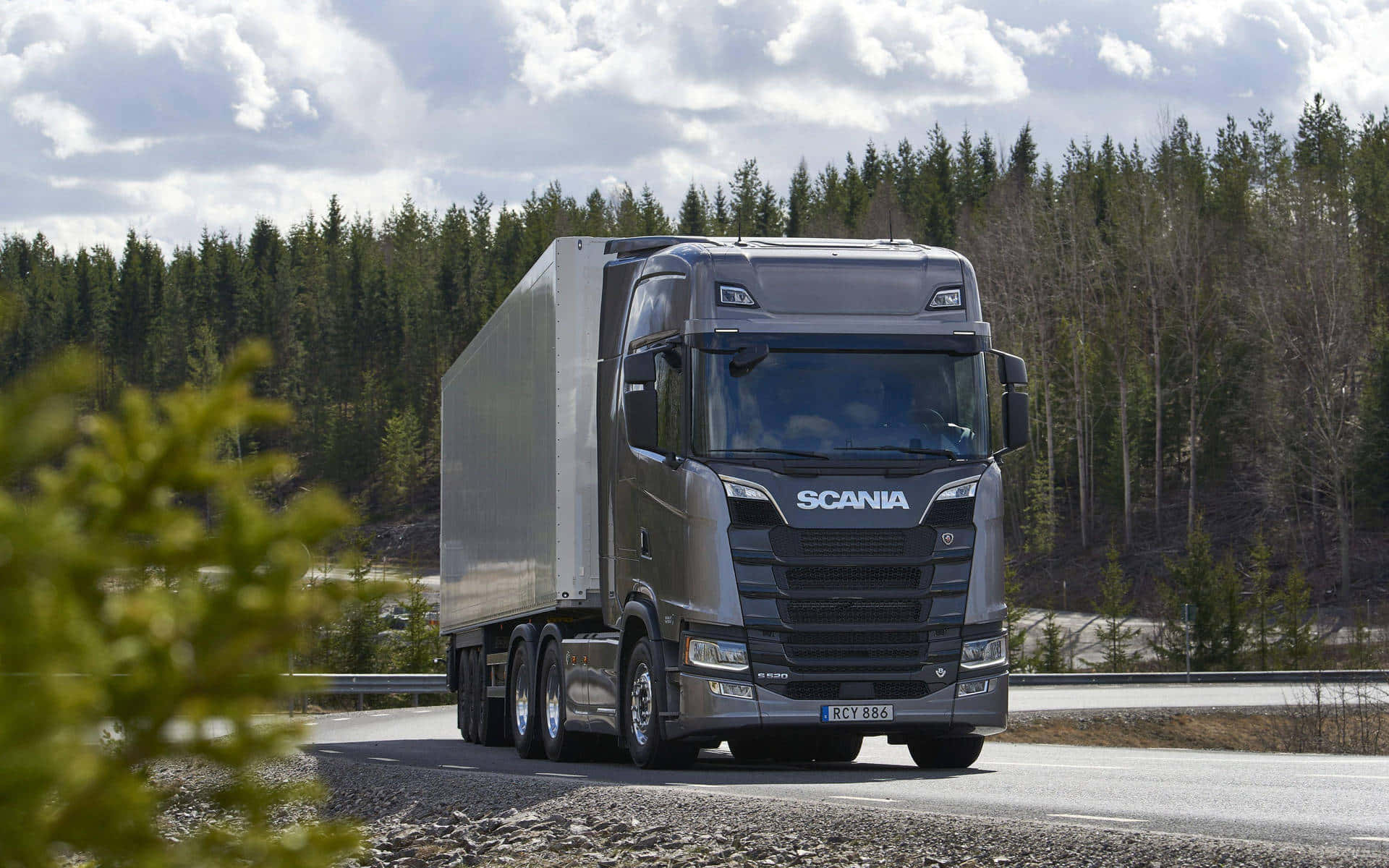 Truck 桌布 壁紙 — 一輛時尚的灰色Scania半掛卡車拖著銀色拖車，在部分多雲的天空下行駛在穿過森林景觀的瀝青路上。