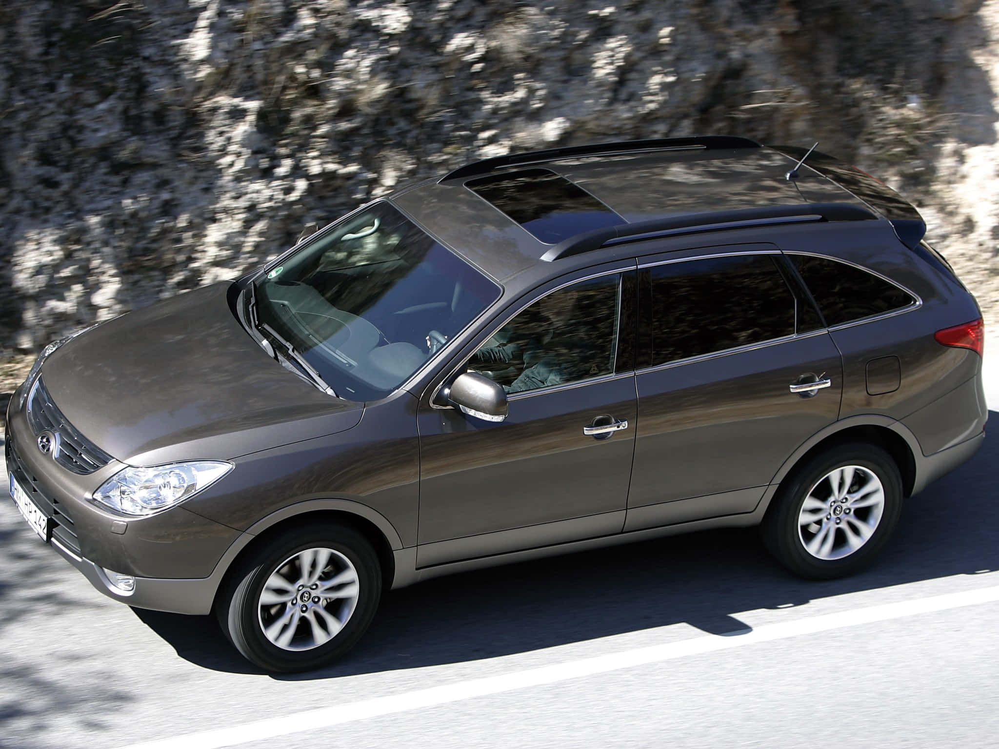Hyundai Veracruz 桌布 壁紙 — 一輛時尚的灰色SUV在陽光明媚的道路上行駛，旁邊是崎嶇的岩石峭壁。