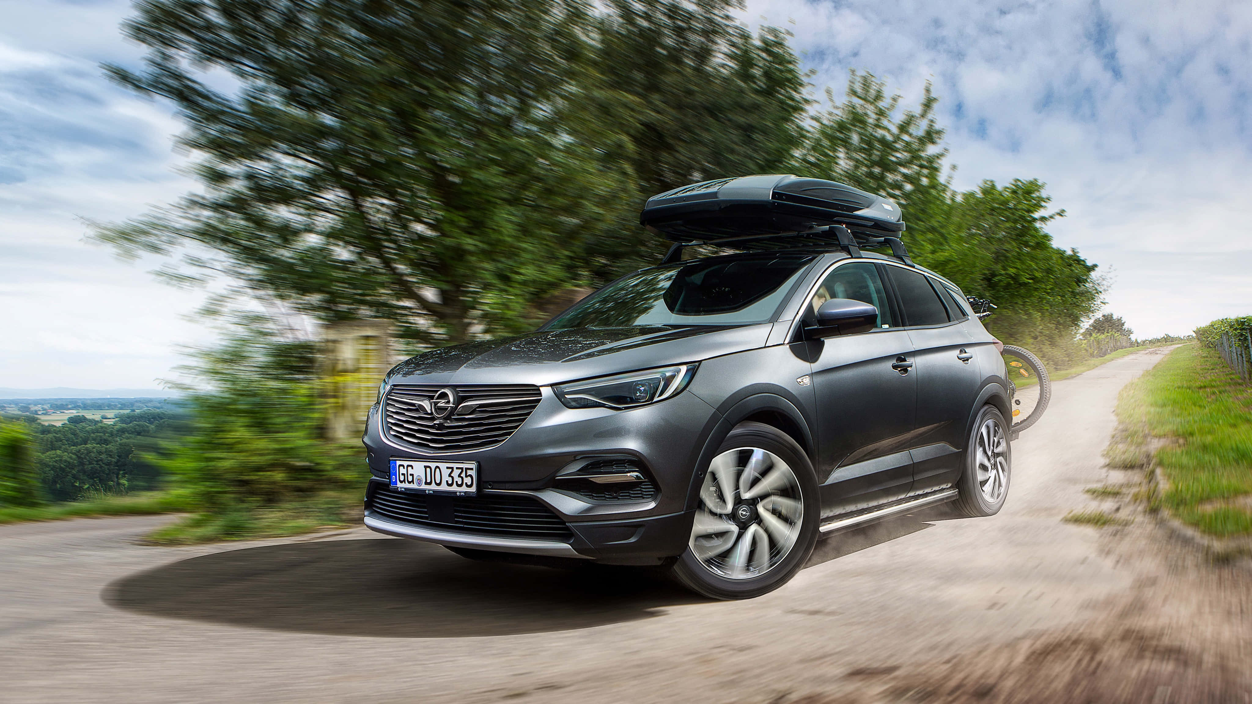 Opel Grandland X 桌布 壁紙 — 一輛時尚的灰色SUV配備車頂行李箱，在部分多雲的天空下，沿著蜿蜒的鄉村道路飛馳，周圍環繞著茂密的綠樹。